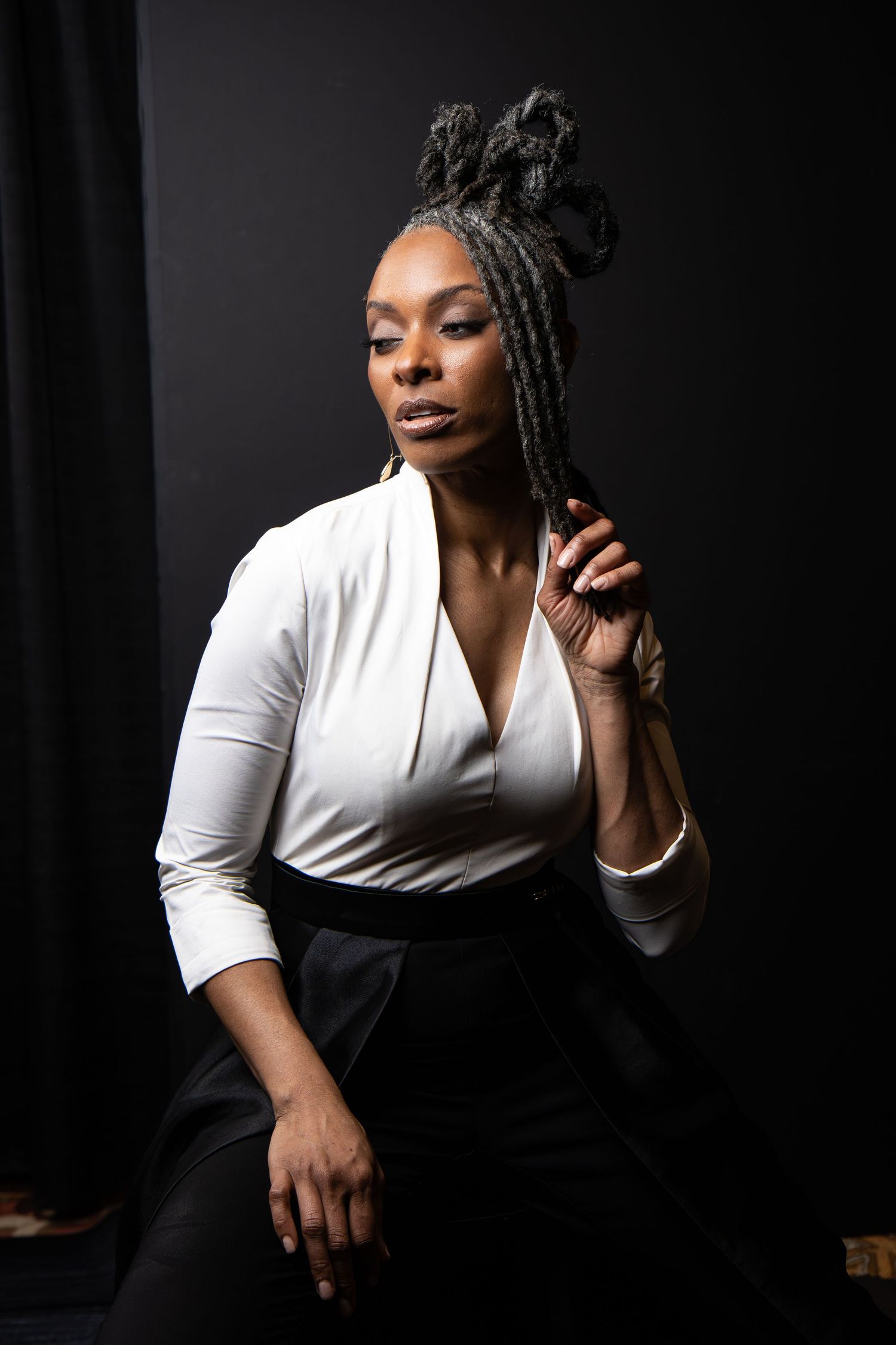 A confident Black woman with braided updo in white top and black pants poses against dark background.