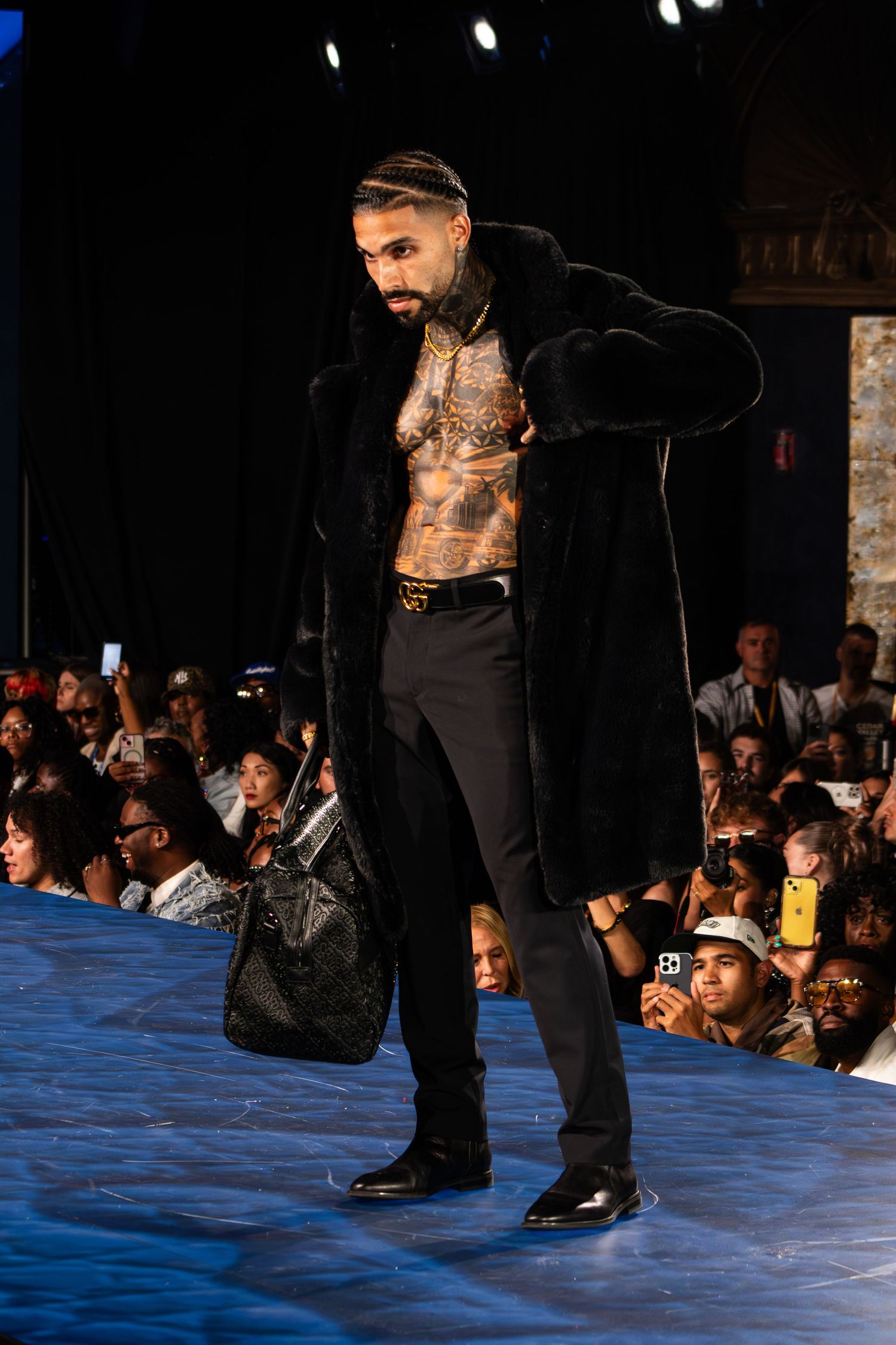 Tattooed male model walks runway in open black fur coat with black pants and bag at fashion show.