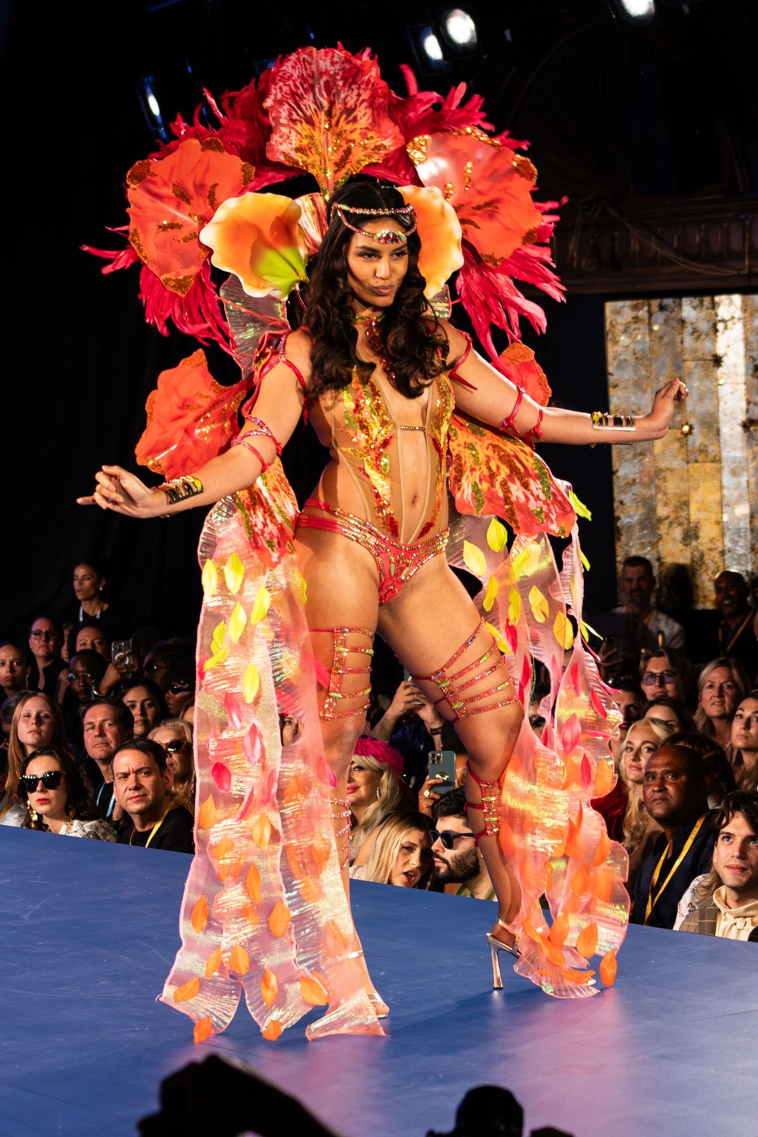 Model walks runway in elaborate orange and red feathered carnival costume with floral headdress.