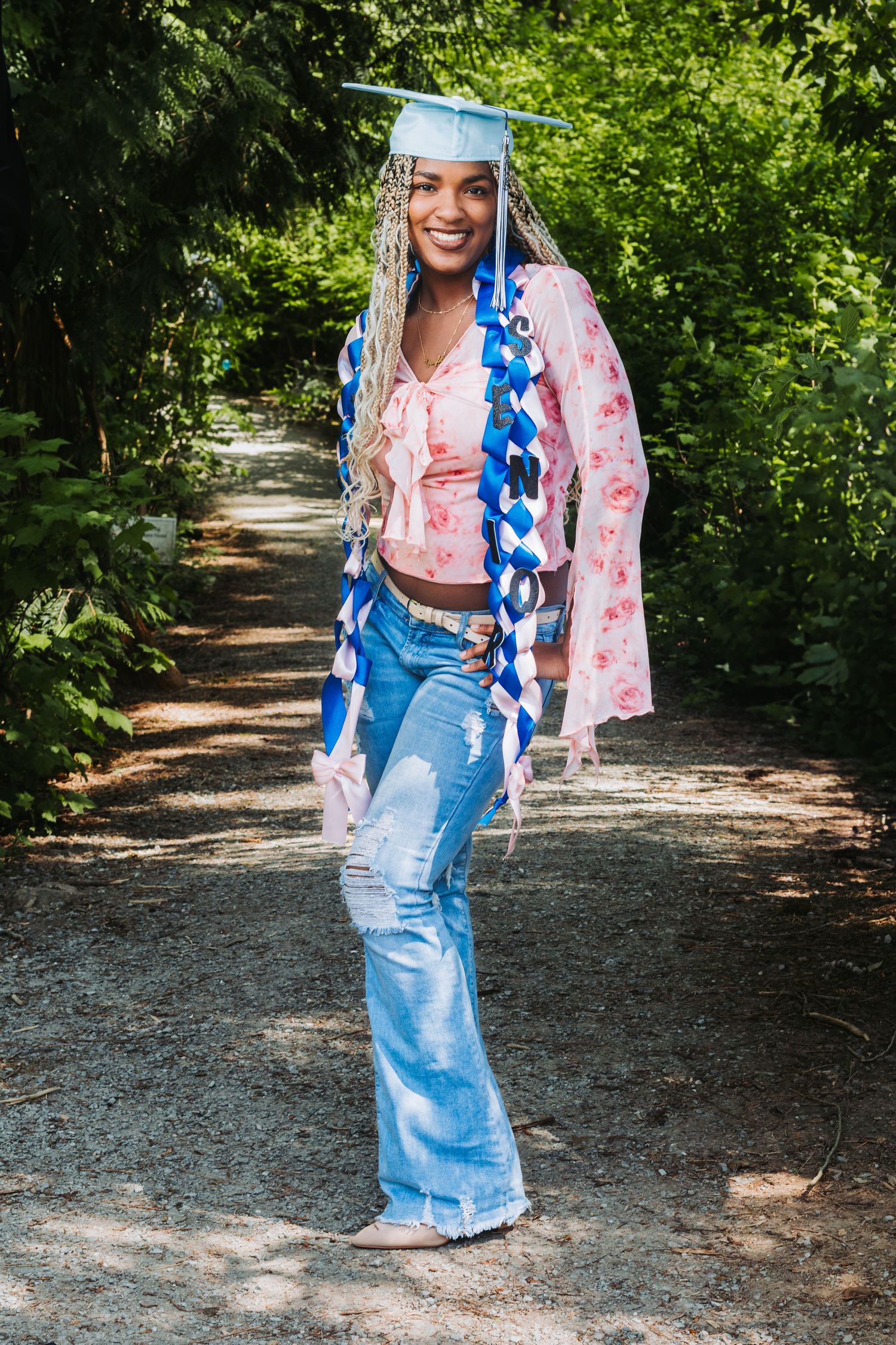 A smiling graduate wearing a blue cap, colorful sashes, floral jacket, and ripped flare jeans poses on a tree-lined path.