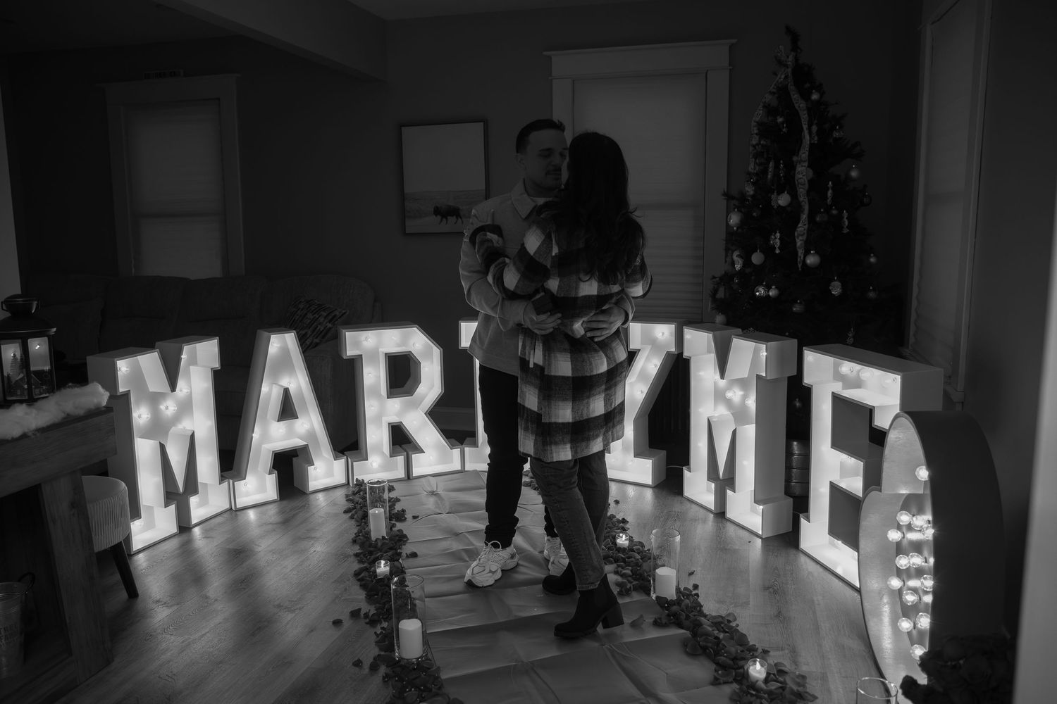Couple embraces in front of illuminated MARRY ME marquee letters near Christmas tree in romantic black and white proposal photo.
