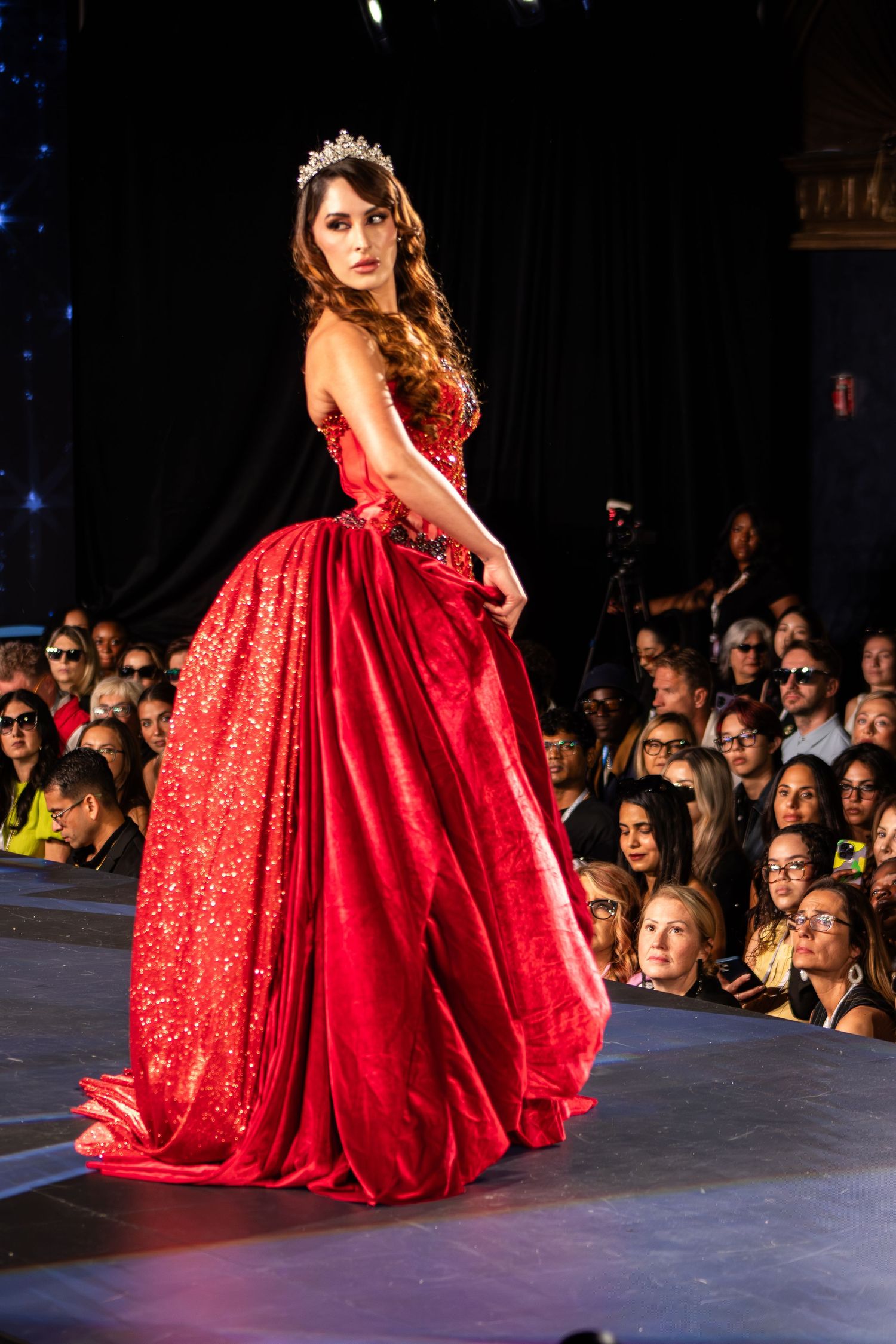 Model in a stunning red ball gown with crystal crown walks the runway at a fashion show.