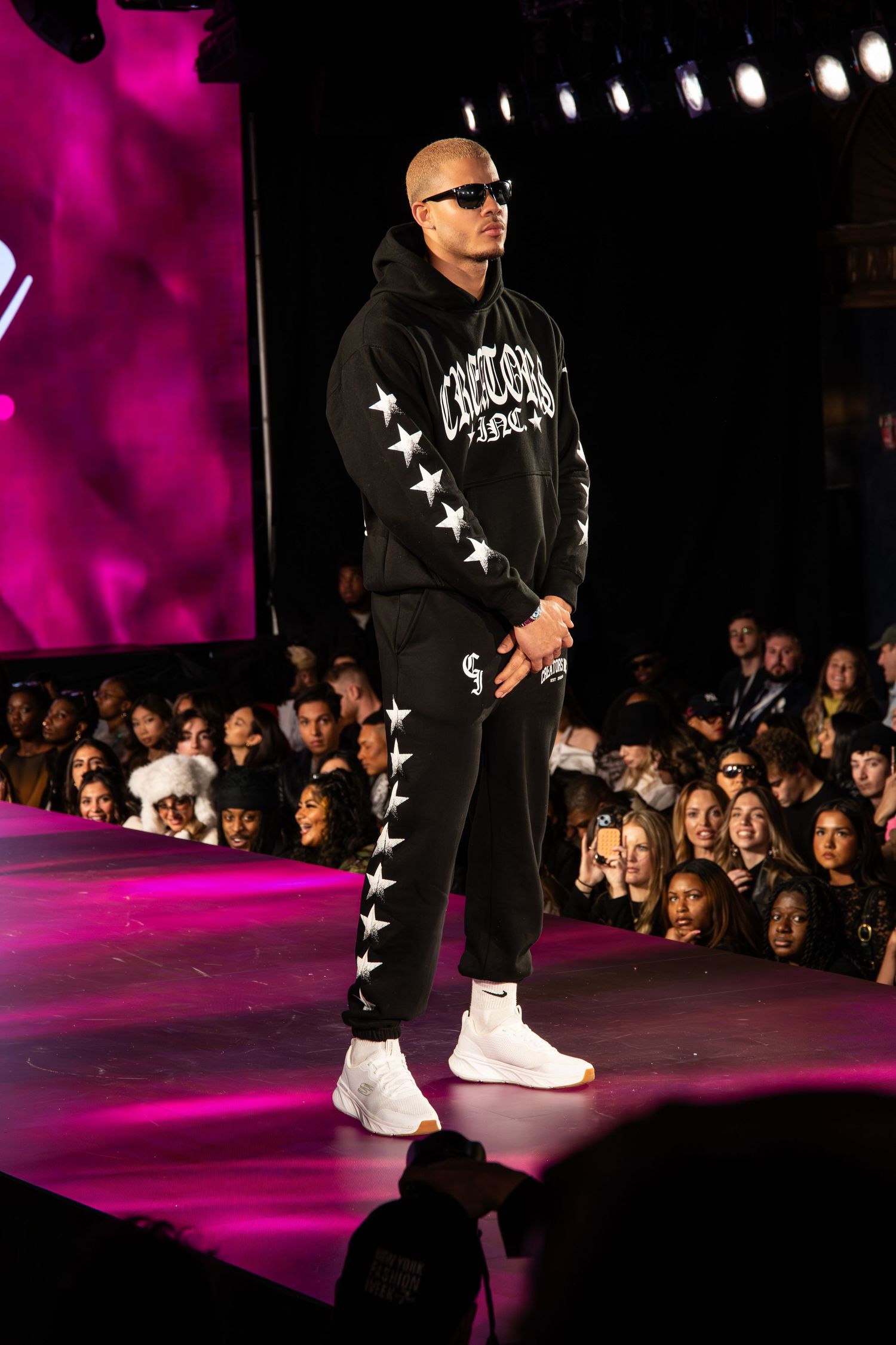 Model walks runway in black streetwear tracksuit with white star details and white sneakers during fashion show.