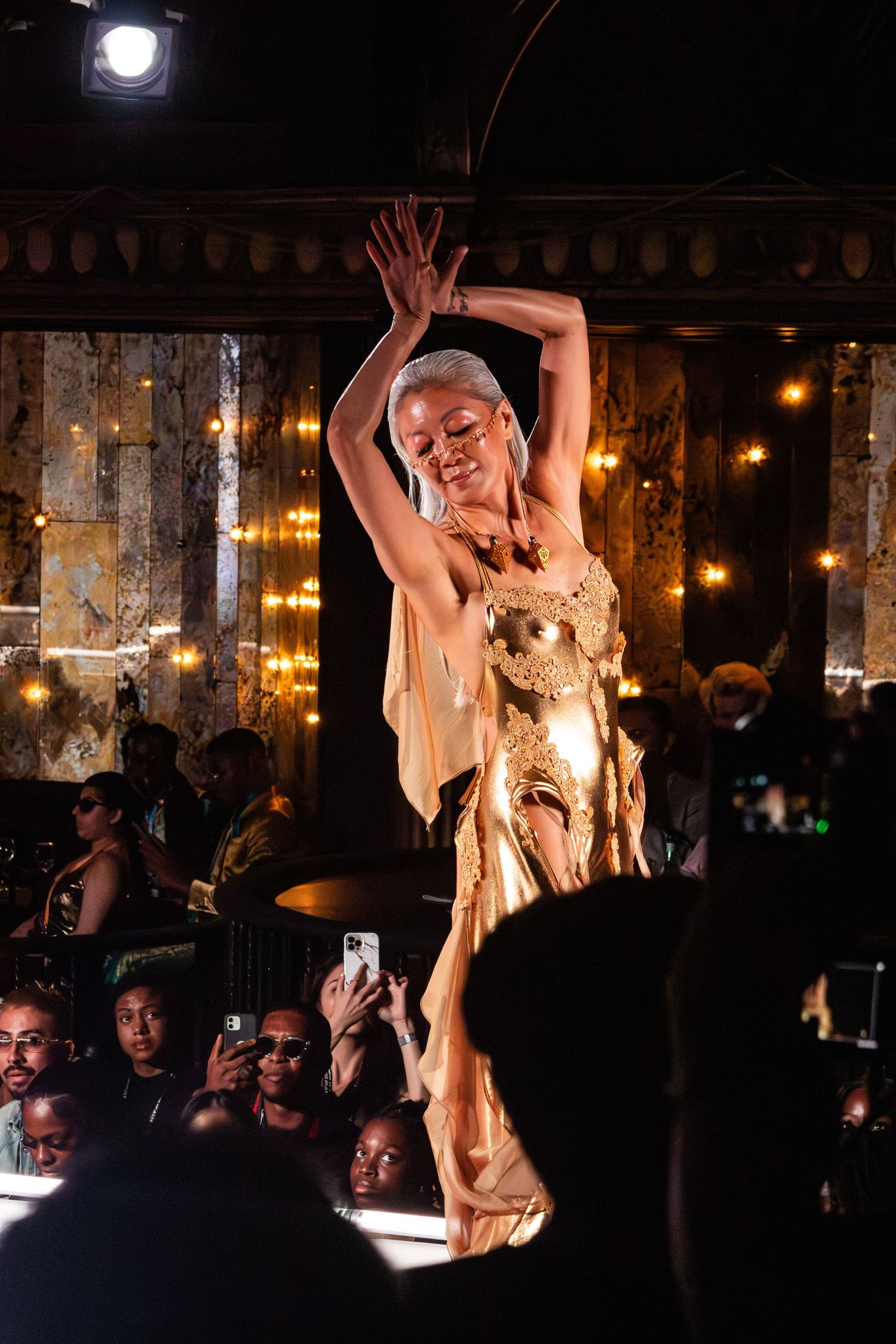 Dancer in gold outfit performs dramatic pose on illuminated stage.
