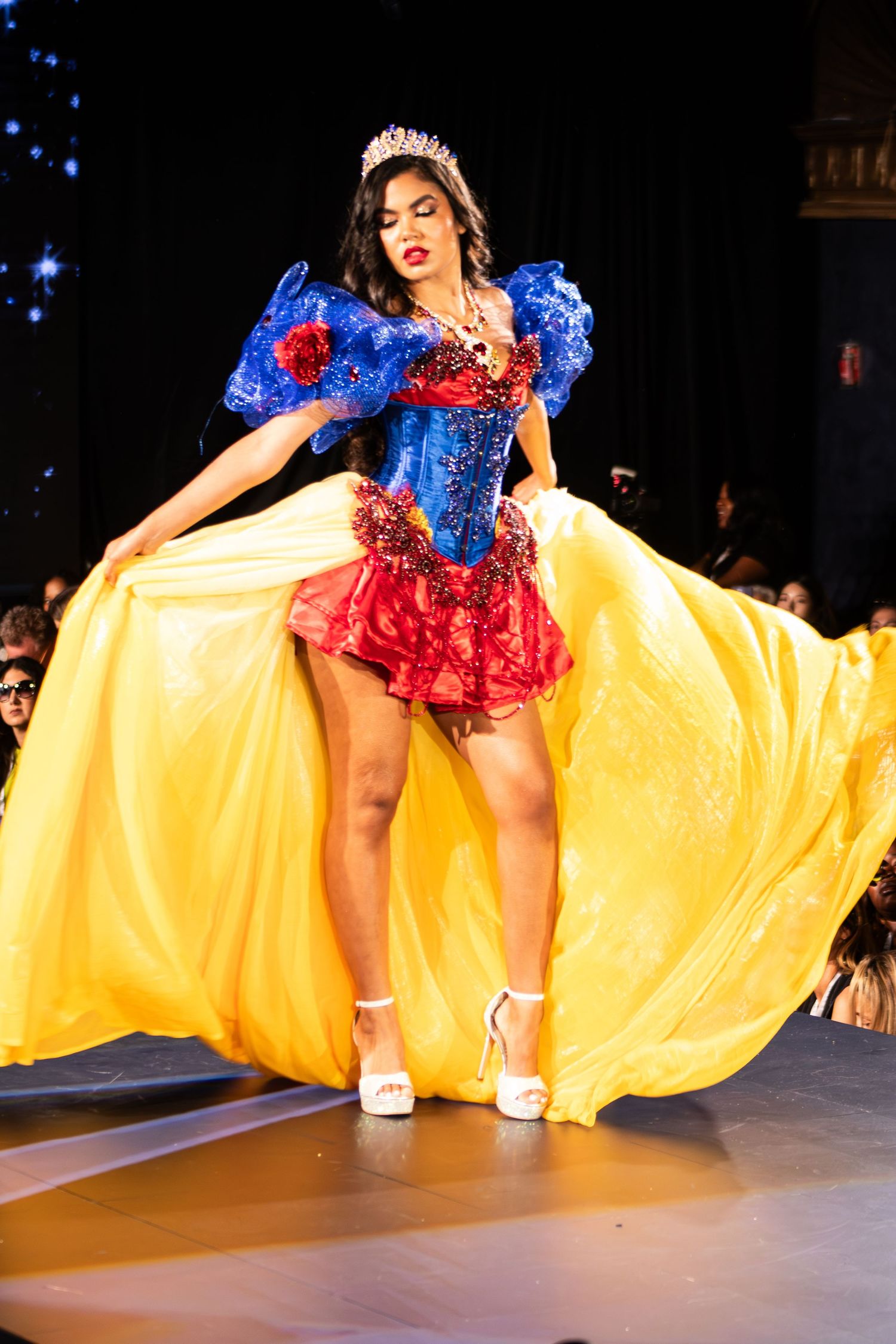 Model showcases Snow White inspired costume with blue bodice and flowing yellow skirt with tiara on runway.