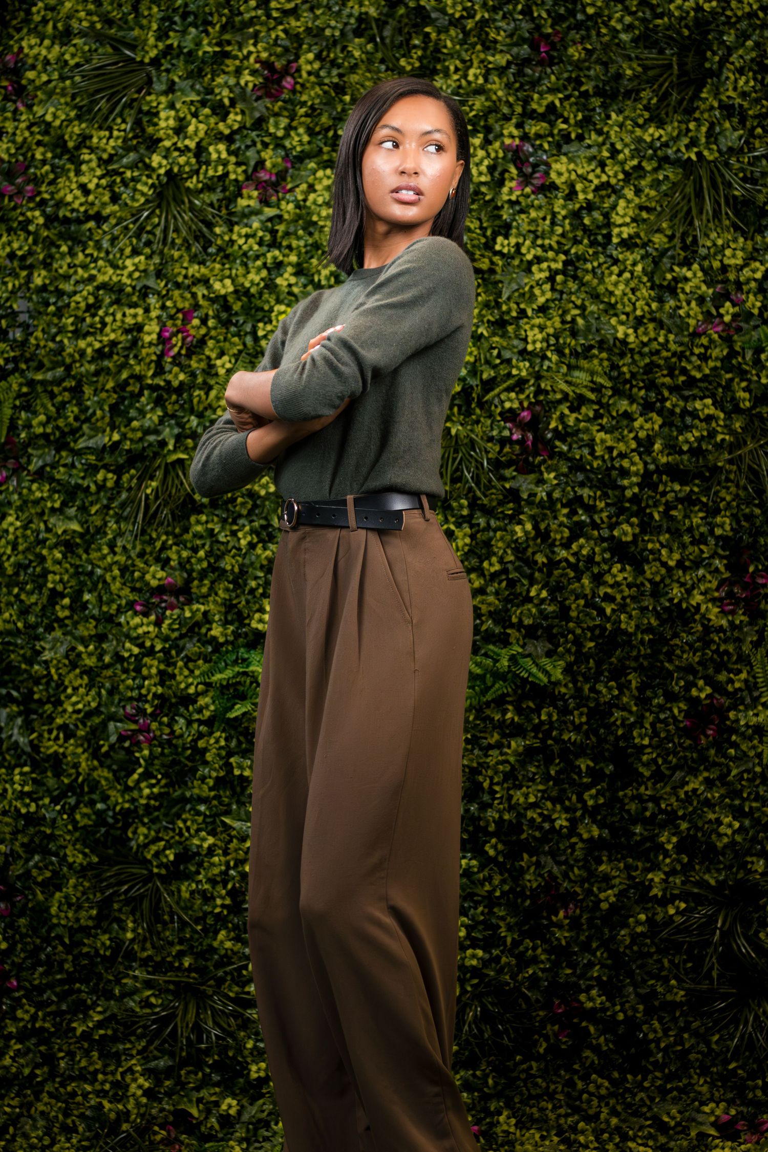 A stylish woman in olive green sweater and brown wide-leg trousers poses confidently against a lush green foliage wall.