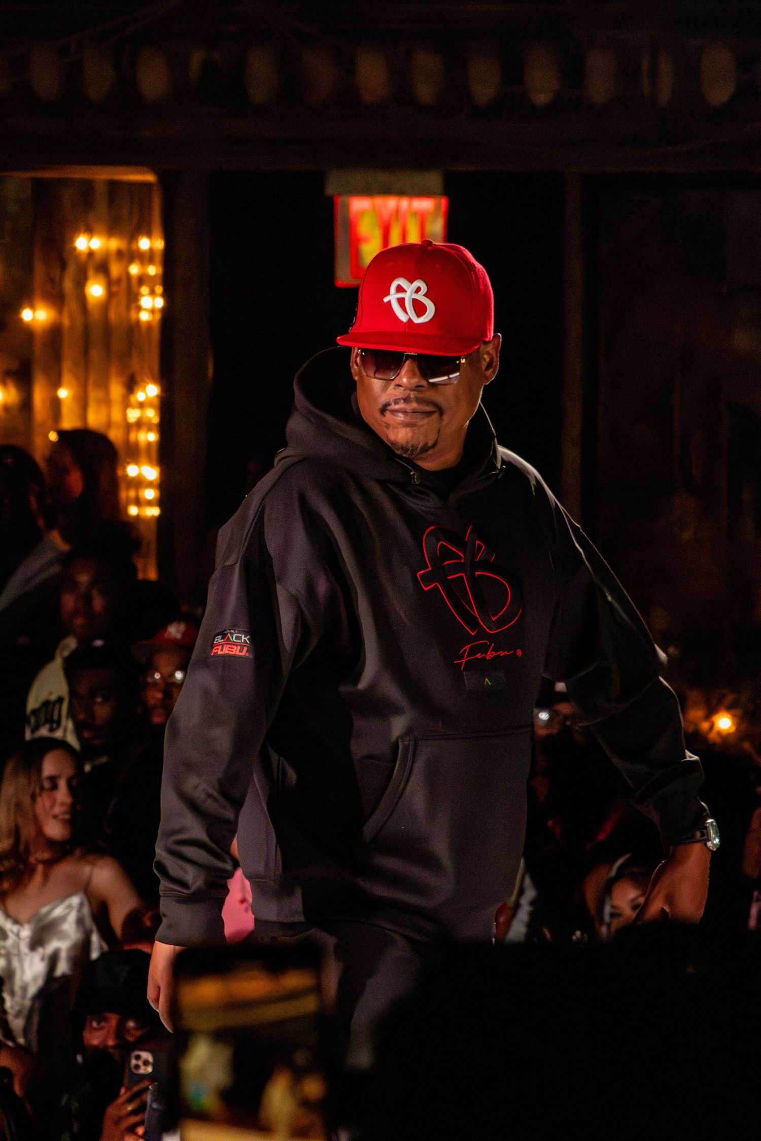 Person wearing red baseball cap and black hoodie performs on dimly lit stage.