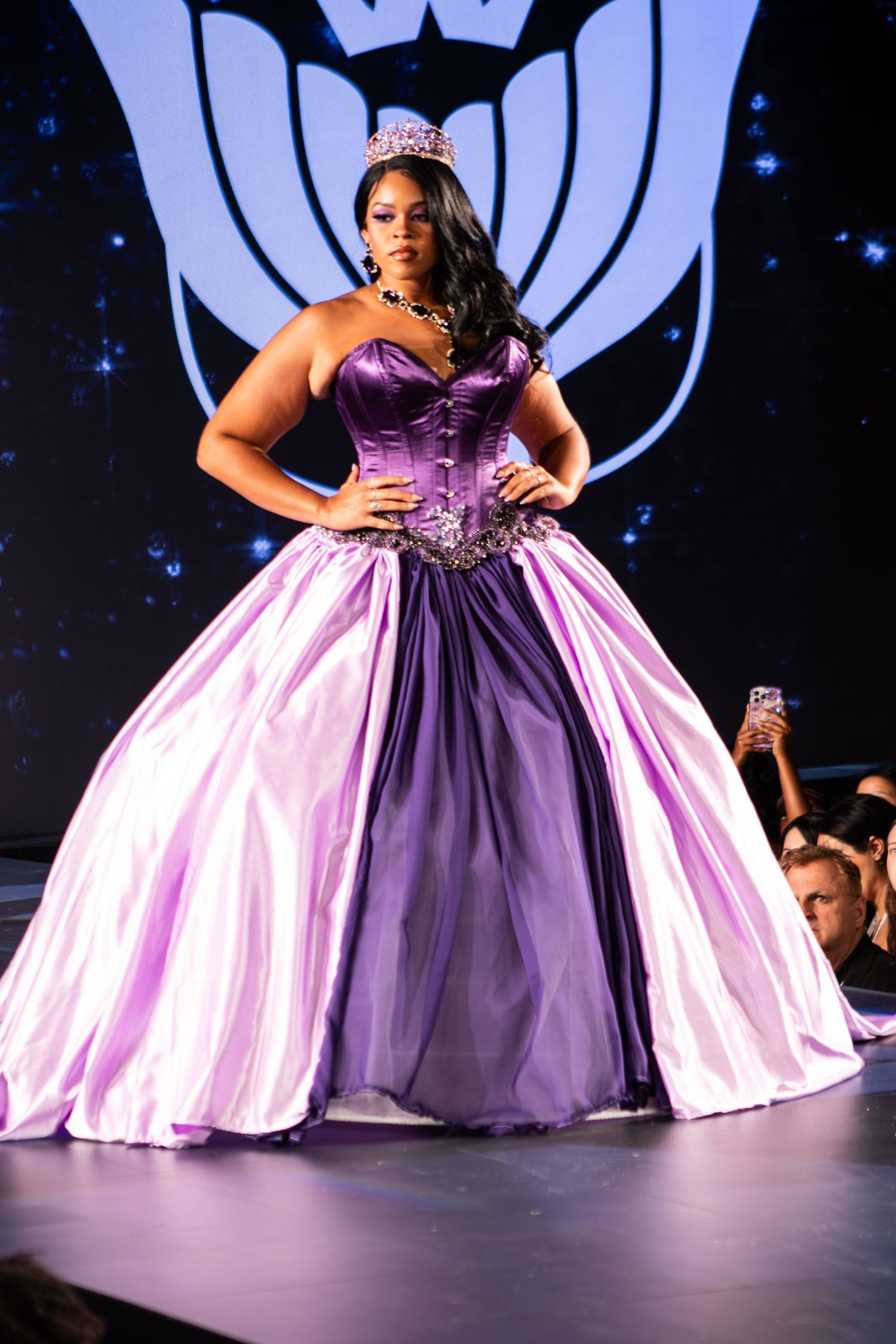 Model walks runway in strapless purple ball gown with tiara and statement necklace at fashion show.