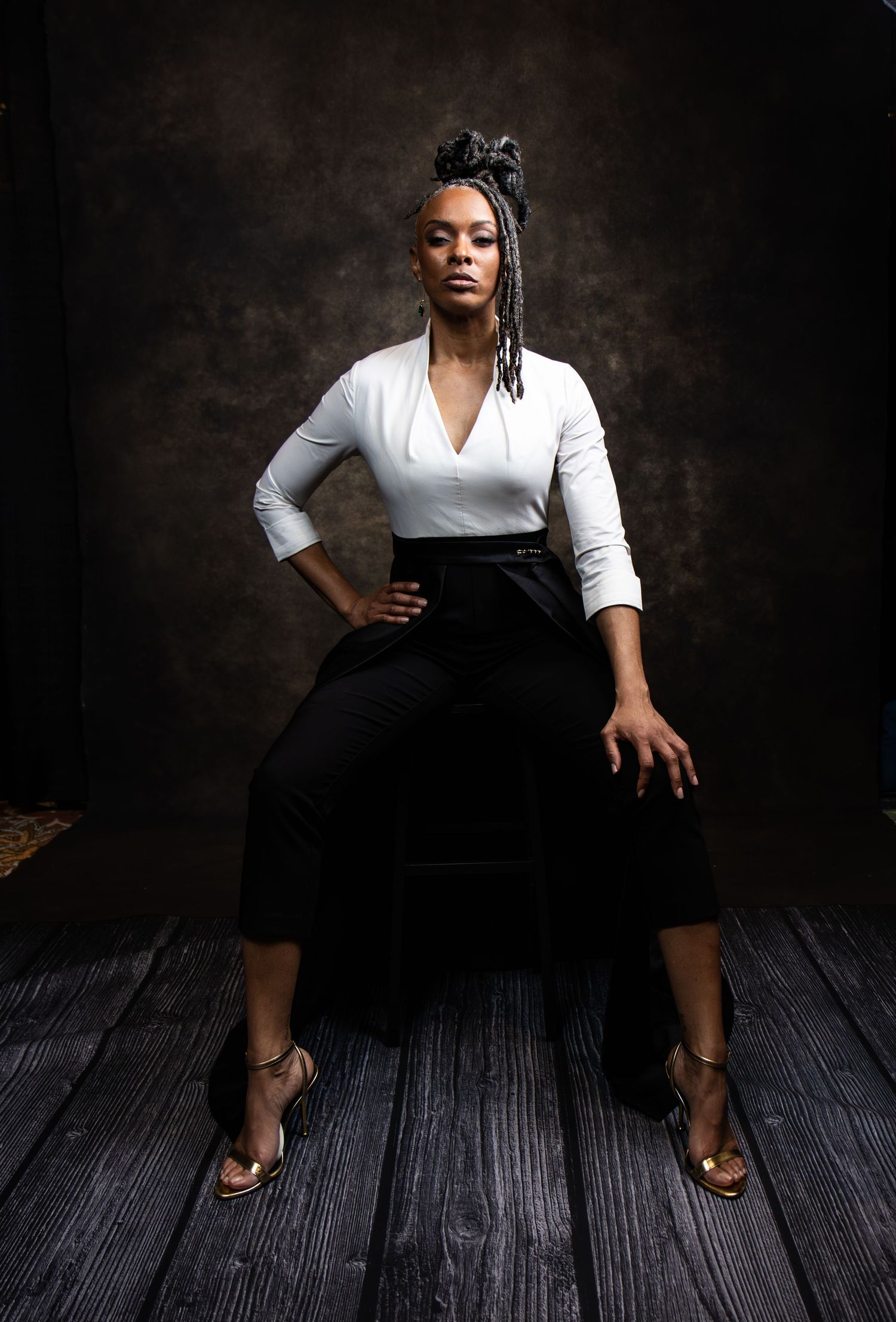 A confident Black woman in a white top and black skirt sits posed in a dramatic studio portrait with moody lighting.