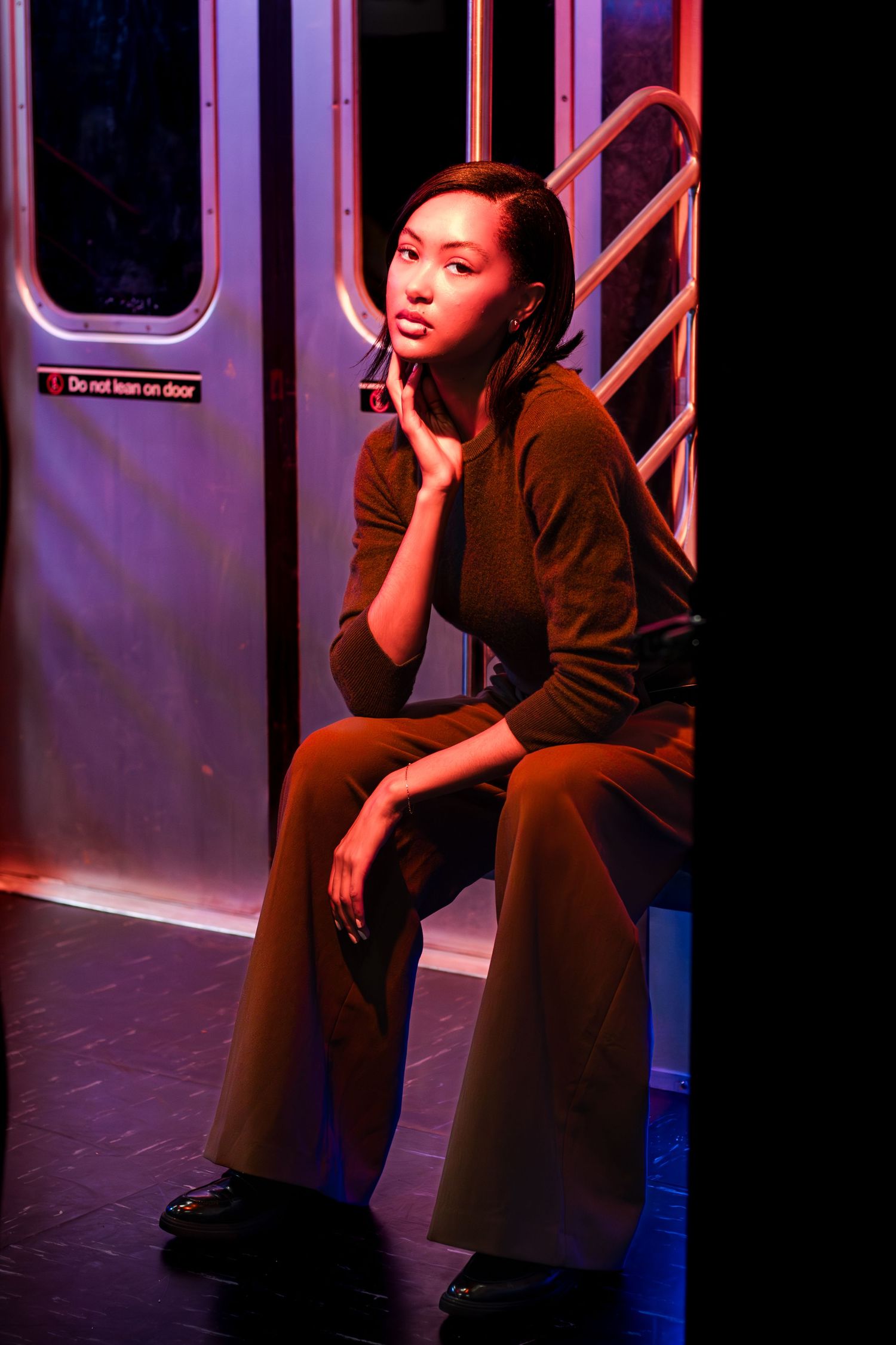 A young woman in an olive green pantsuit sits pensively on a subway car bench, posed against colorful ambient lighting.