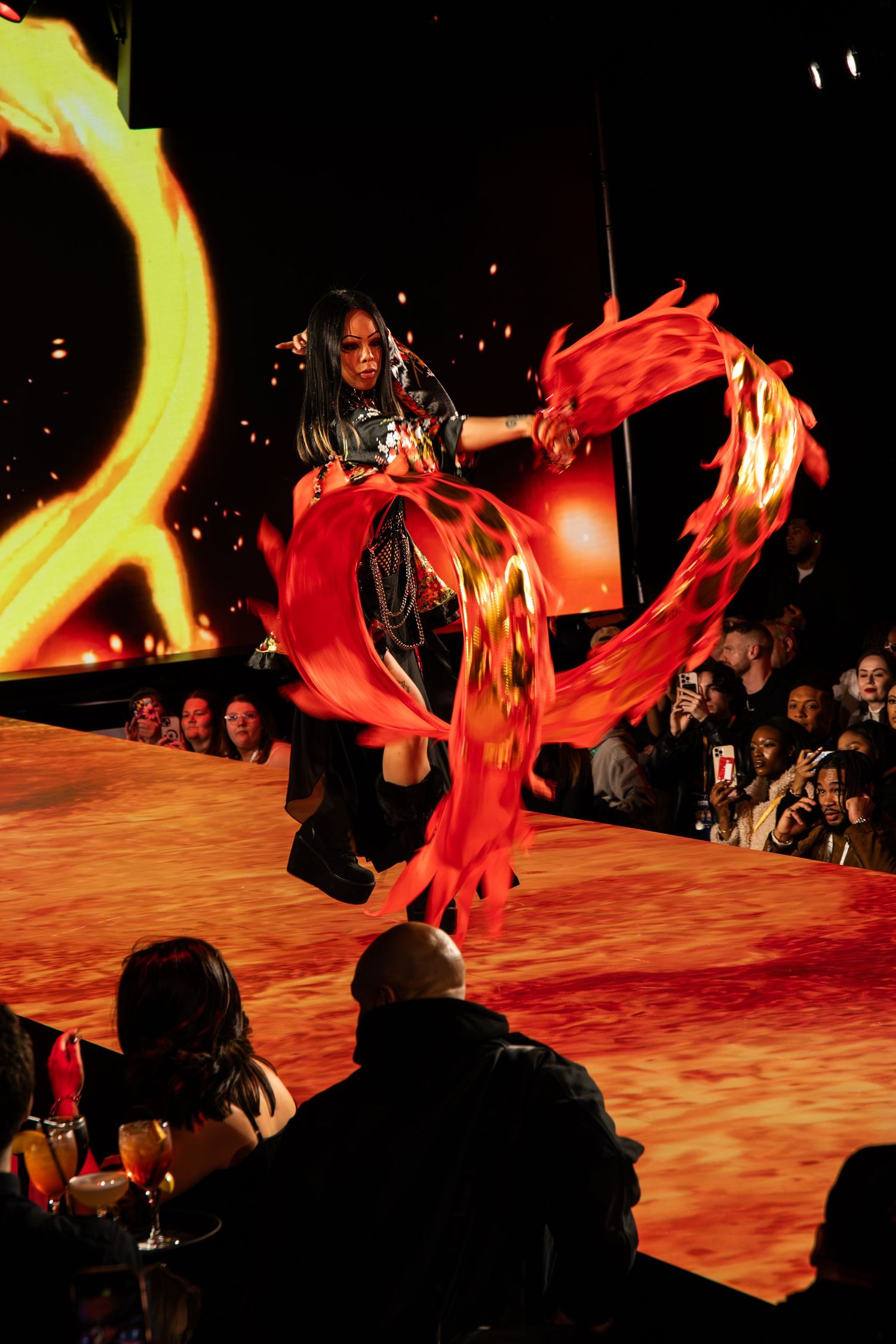 A dancer performs with flowing red fabric under dramatic lighting on a stage with audience silhouettes visible below.
