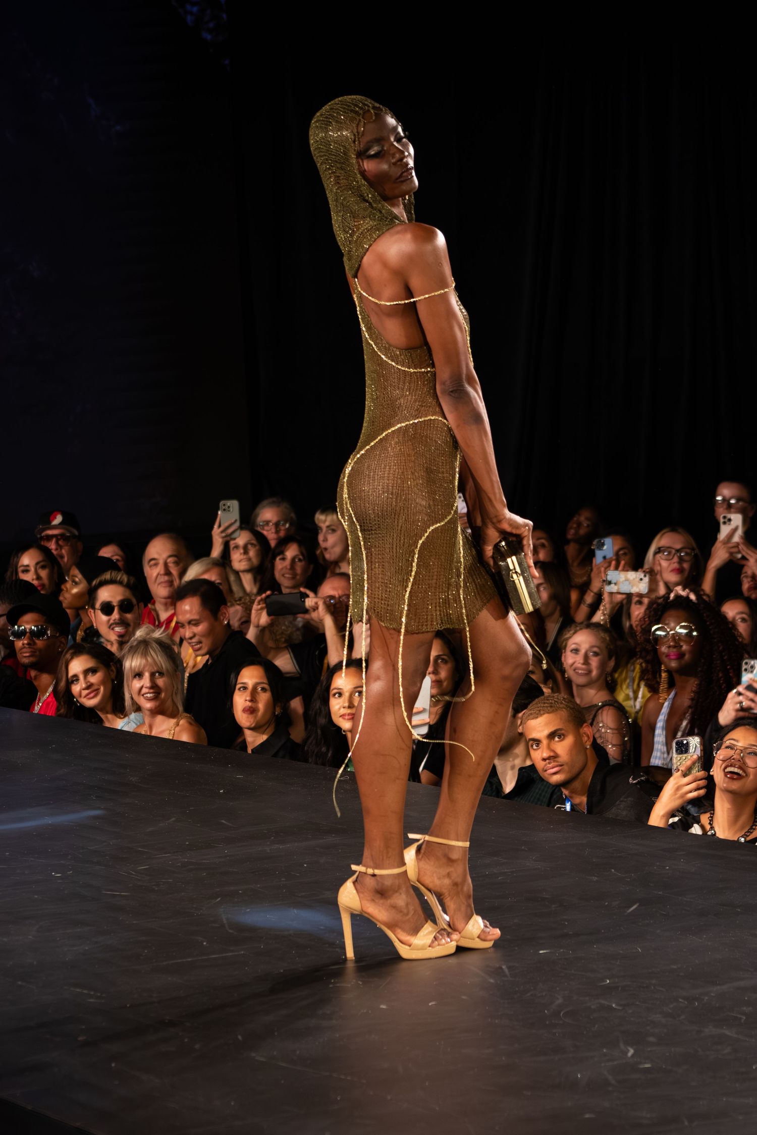 Female model poses on runway in gold beaded mini dress with matching headpiece and strappy heels.