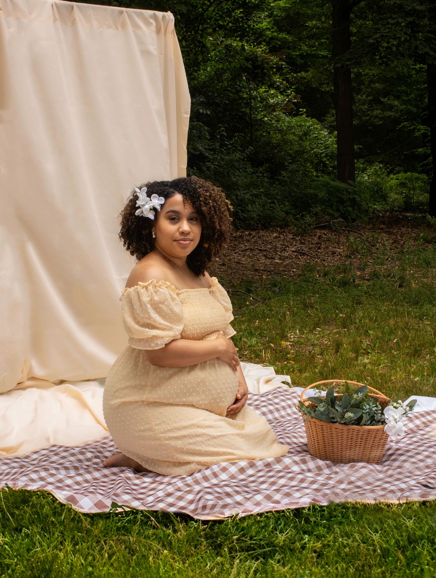 Maternity picnic scene with cream dress and wicker basket on checkered blanket.