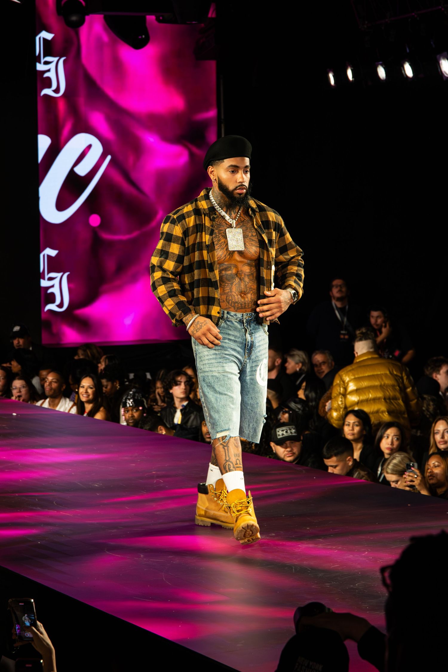 Model walks runway in plaid flannel shirt and ripped jeans during fashion show with pink lighting and backdrop.