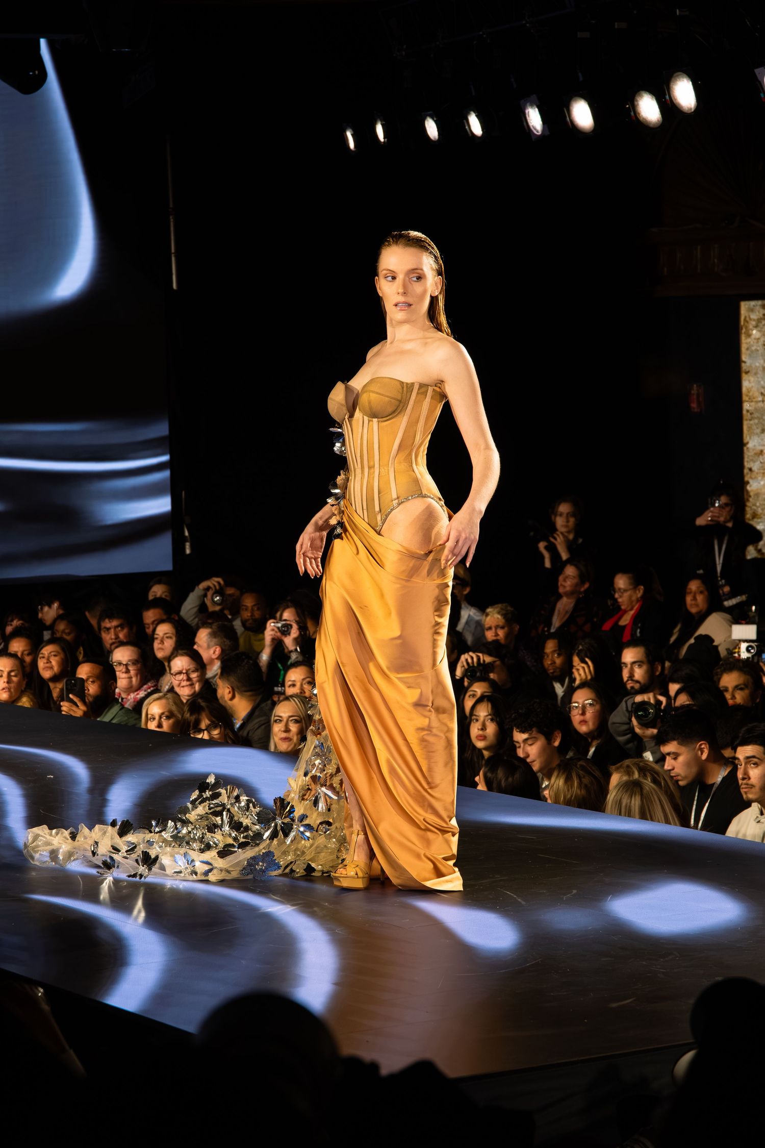 Model walks runway in golden silk evening gown with corset detail during fashion show with dramatic lighting effects.
