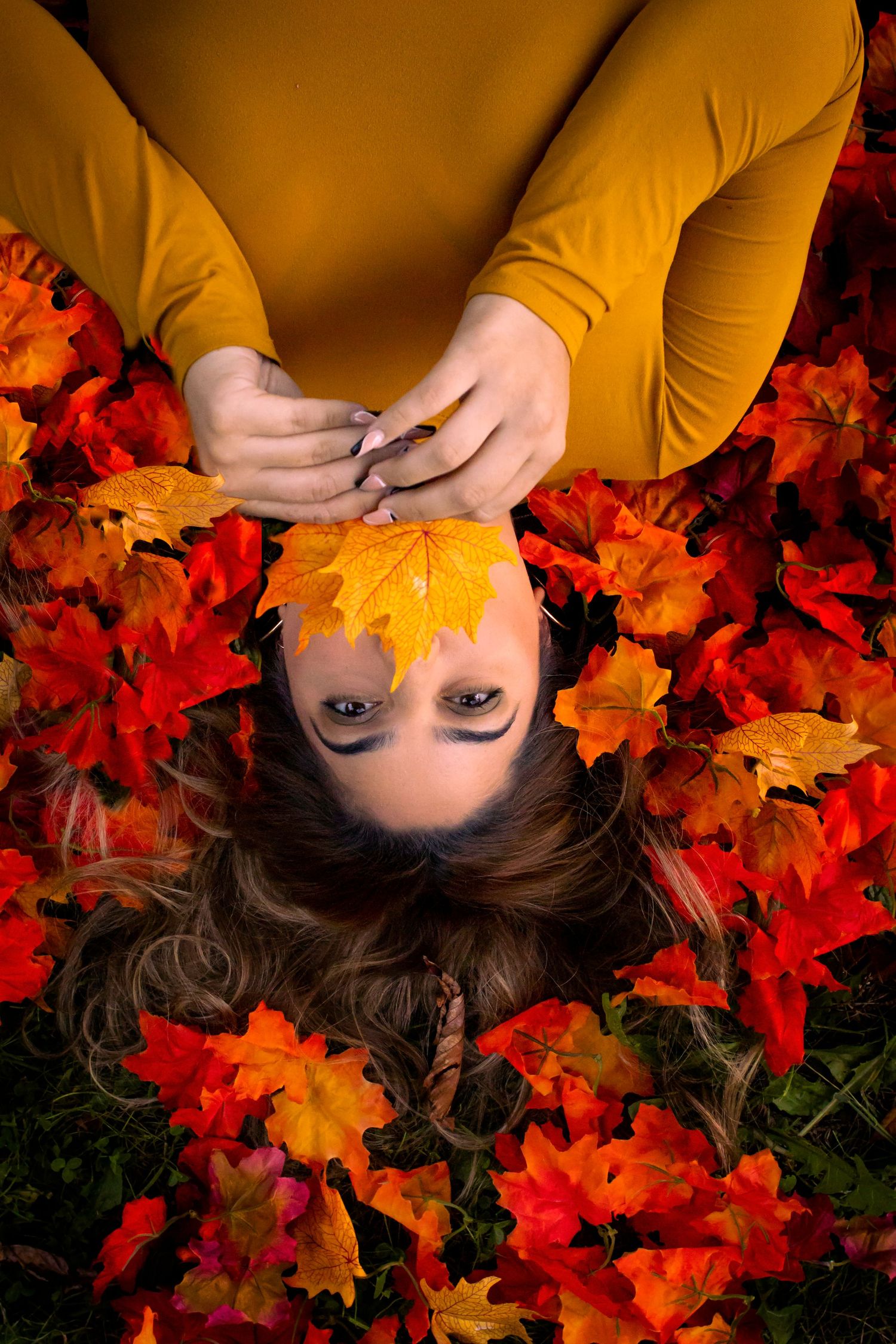 Person lying in vibrant red and orange autumn maple leaves creating seasonal portrait composition.