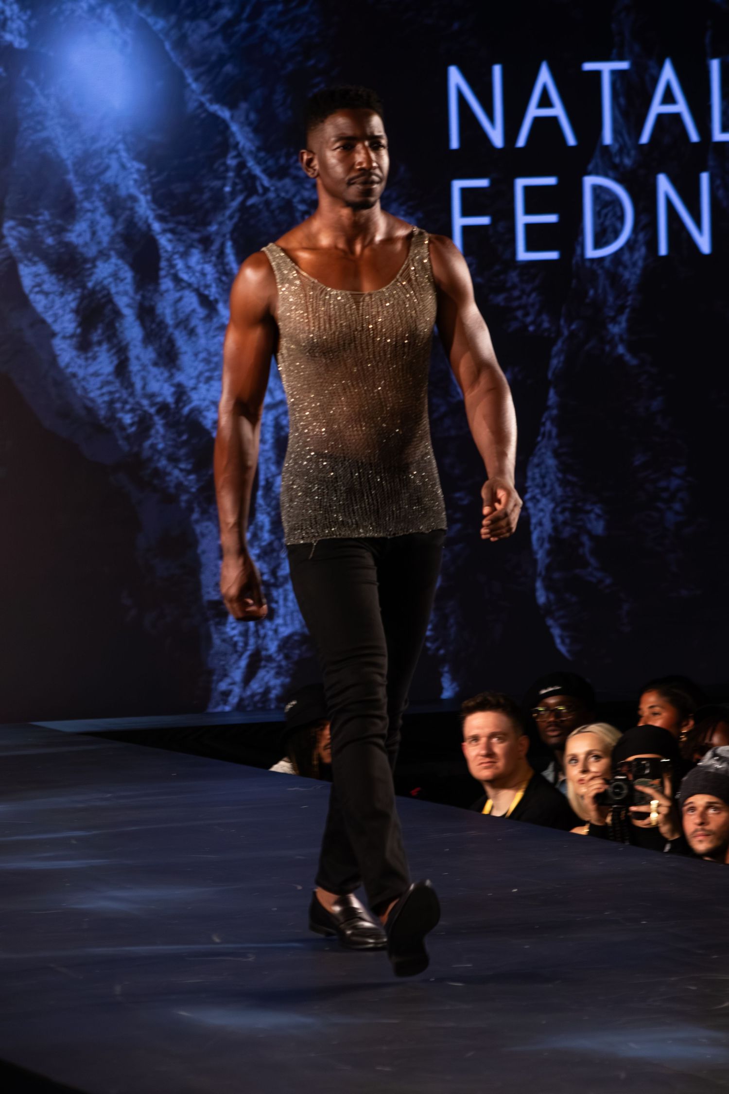 Male model walks runway in sheer embellished tank top and black pants at Natalia Fedner fashion show.
