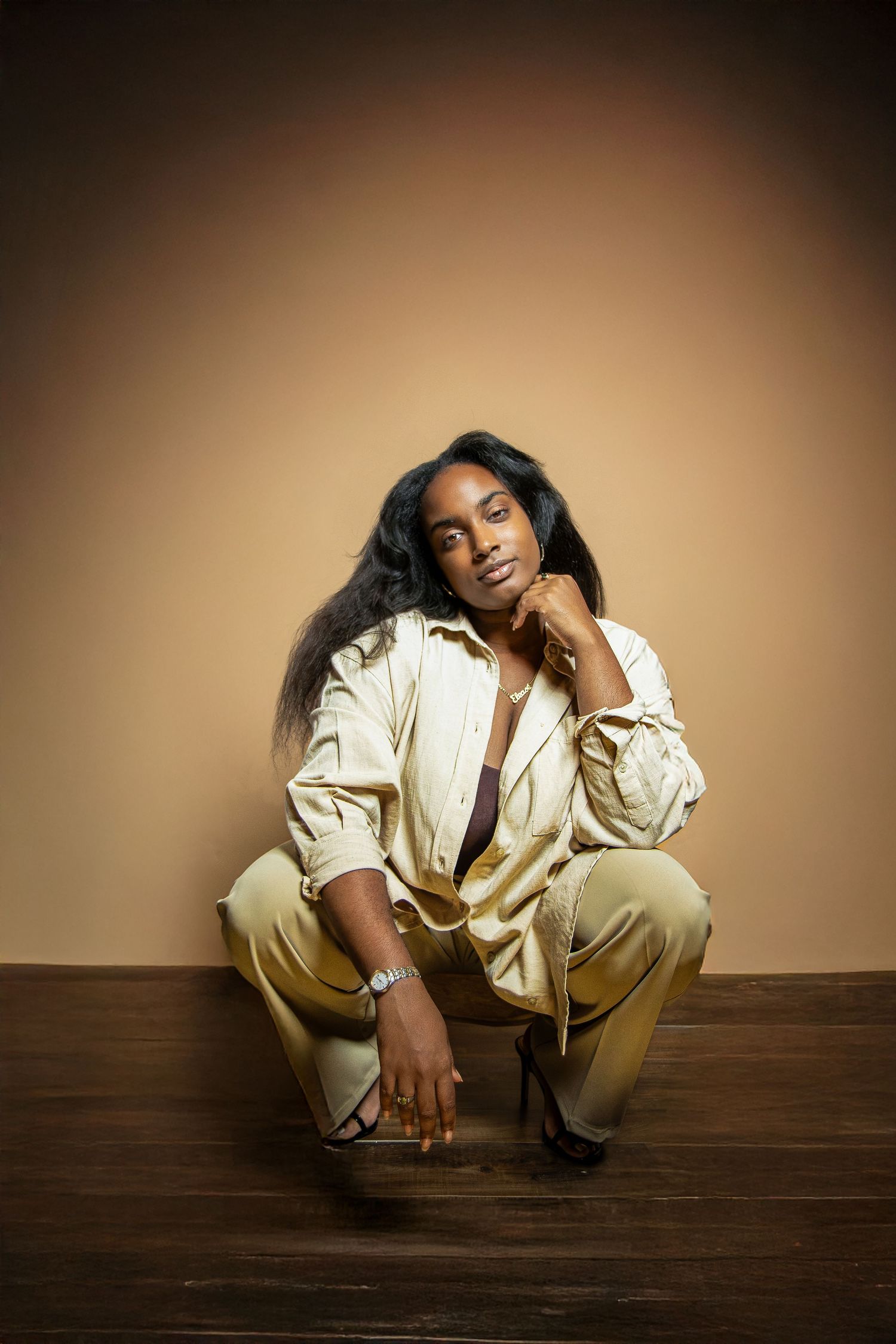 A young Black woman in a cream outfit sits gracefully on a wooden floor against a warm beige background.