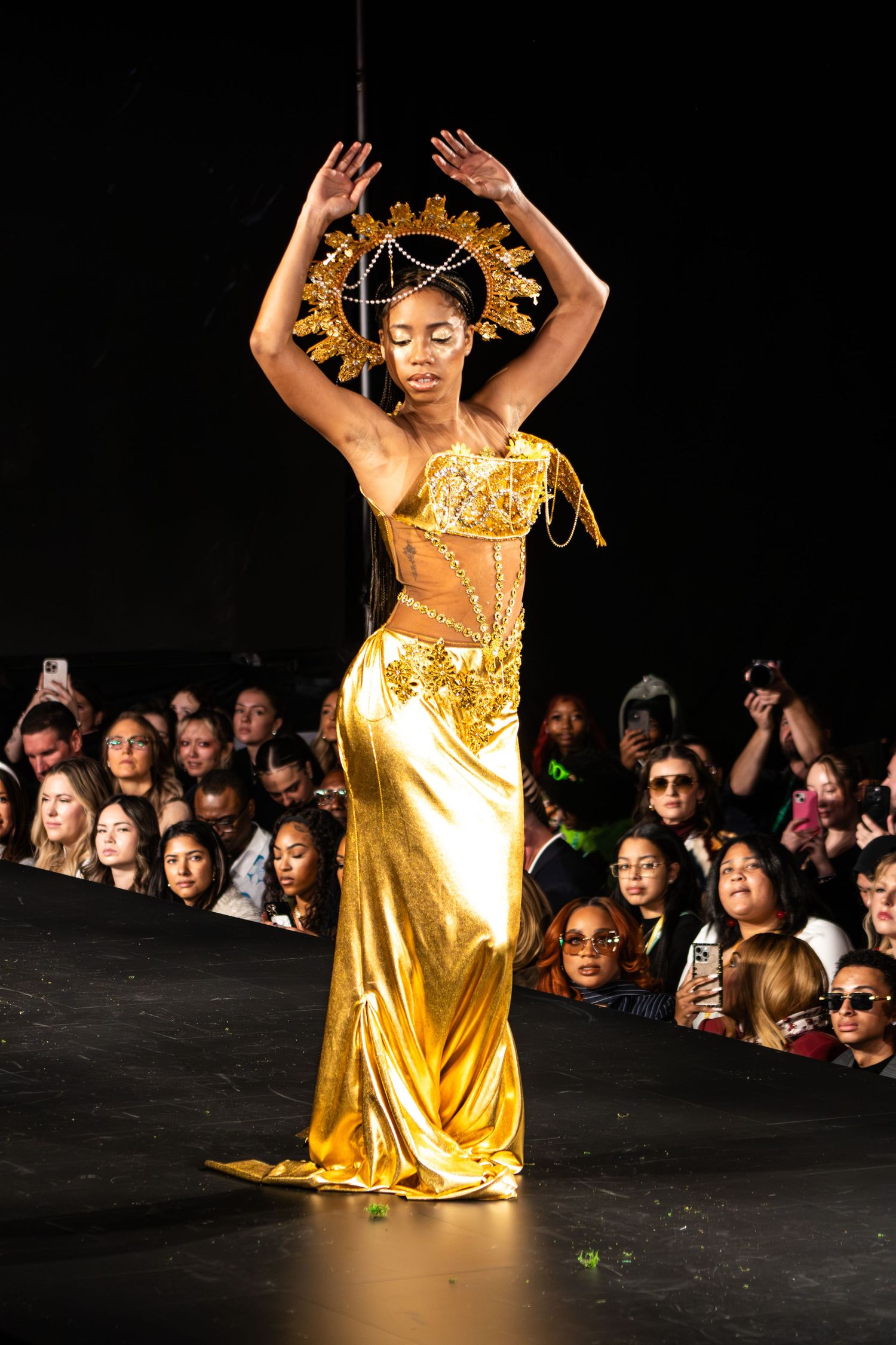 A dancer in a shimmering gold dress and ornate headpiece performs on stage in front of an audience.