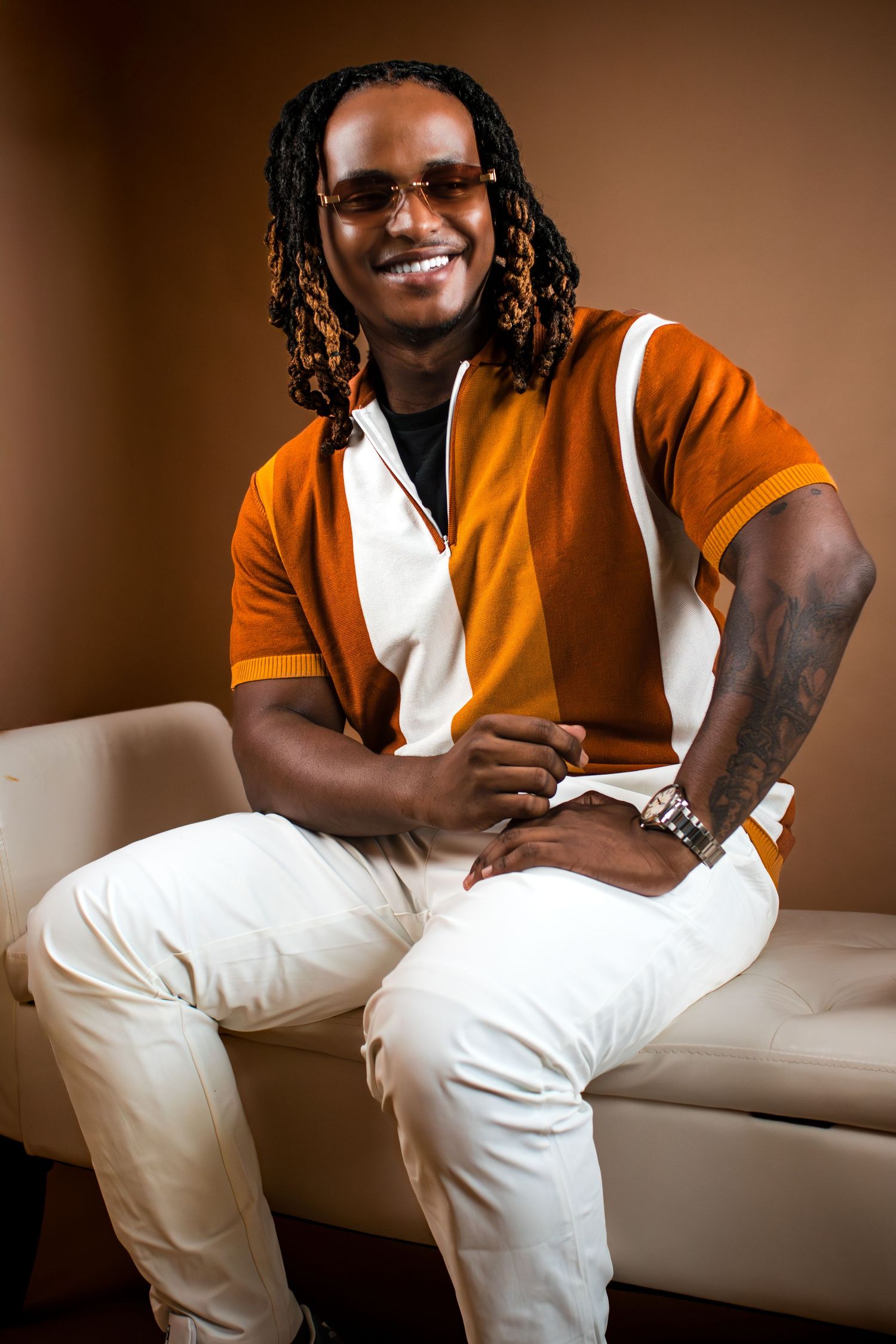 Smiling person in orange and white shirt with dreadlocks sitting casually on white couch against brown backdrop.