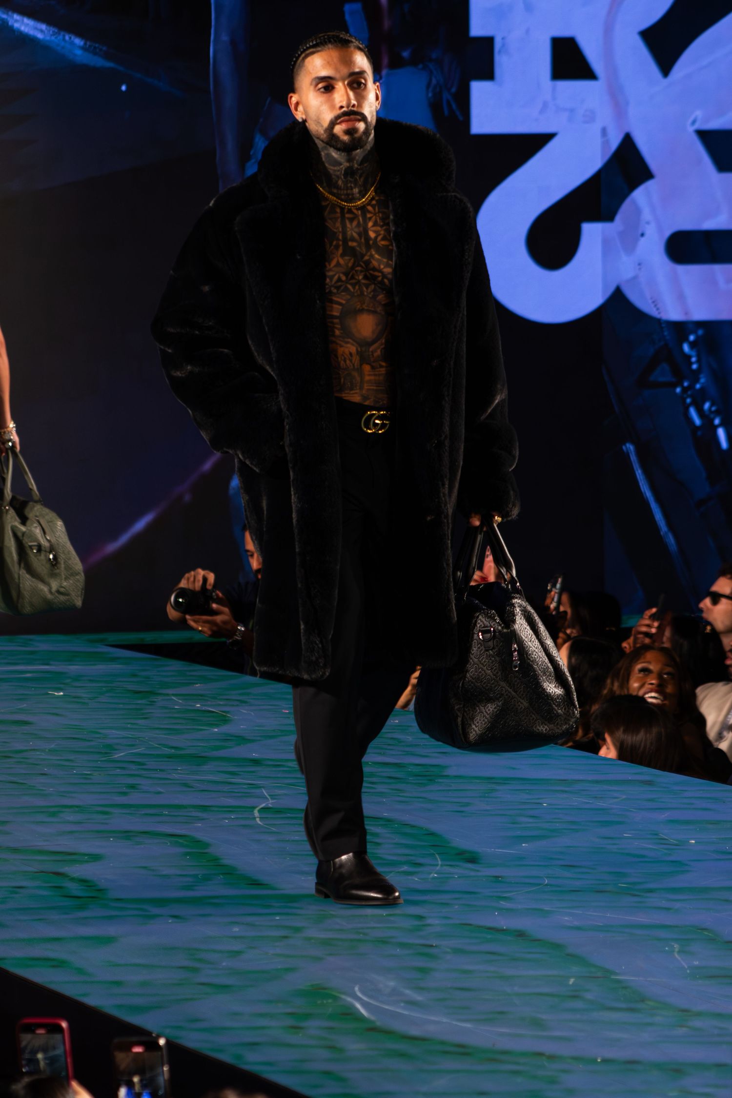 Tattooed male model walks runway in black fur coat with Gucci belt and carrying black bag at fashion show.