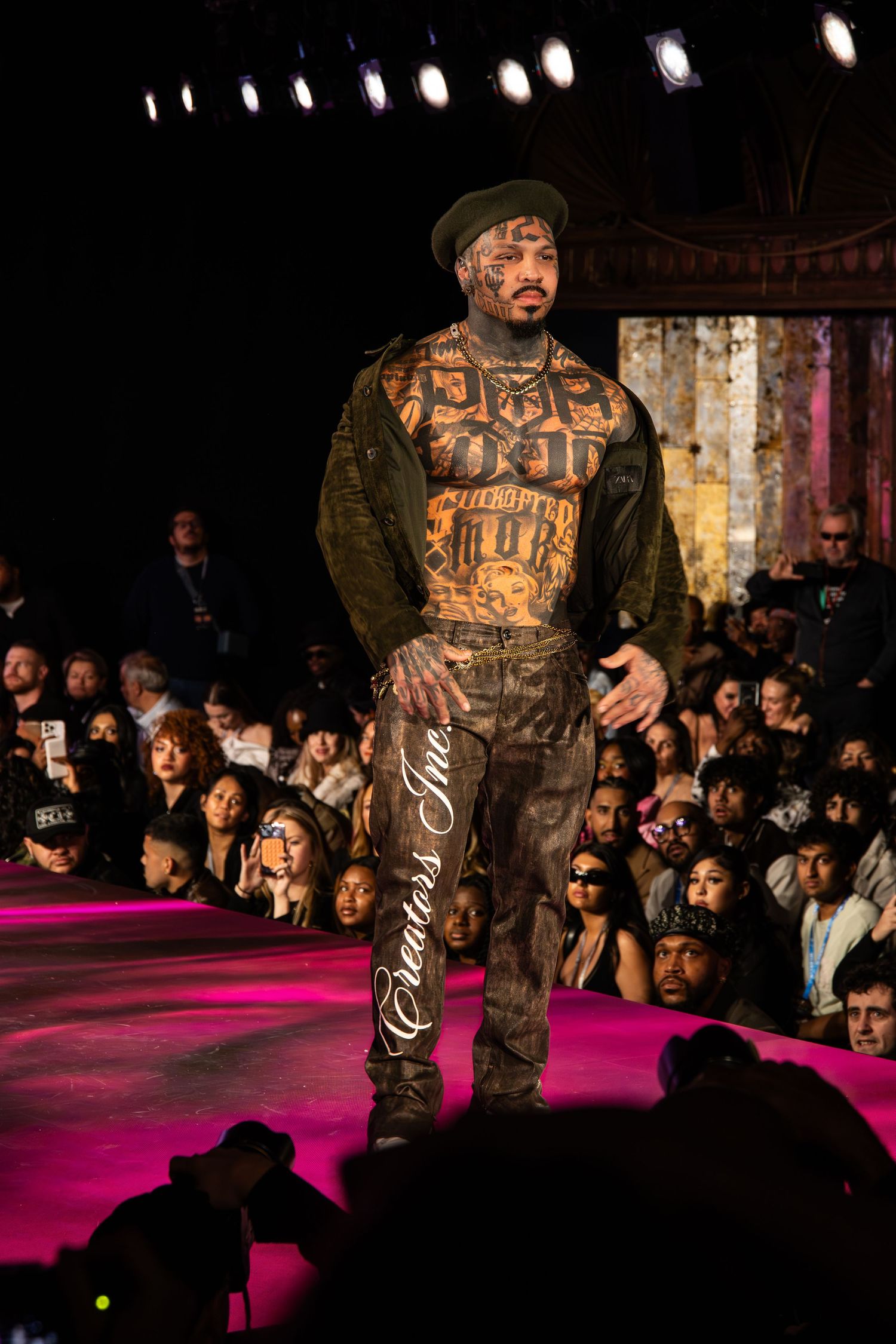 A person walks on a fashion runway wearing an urban streetwear outfit with brown clothing under dramatic stage lighting.