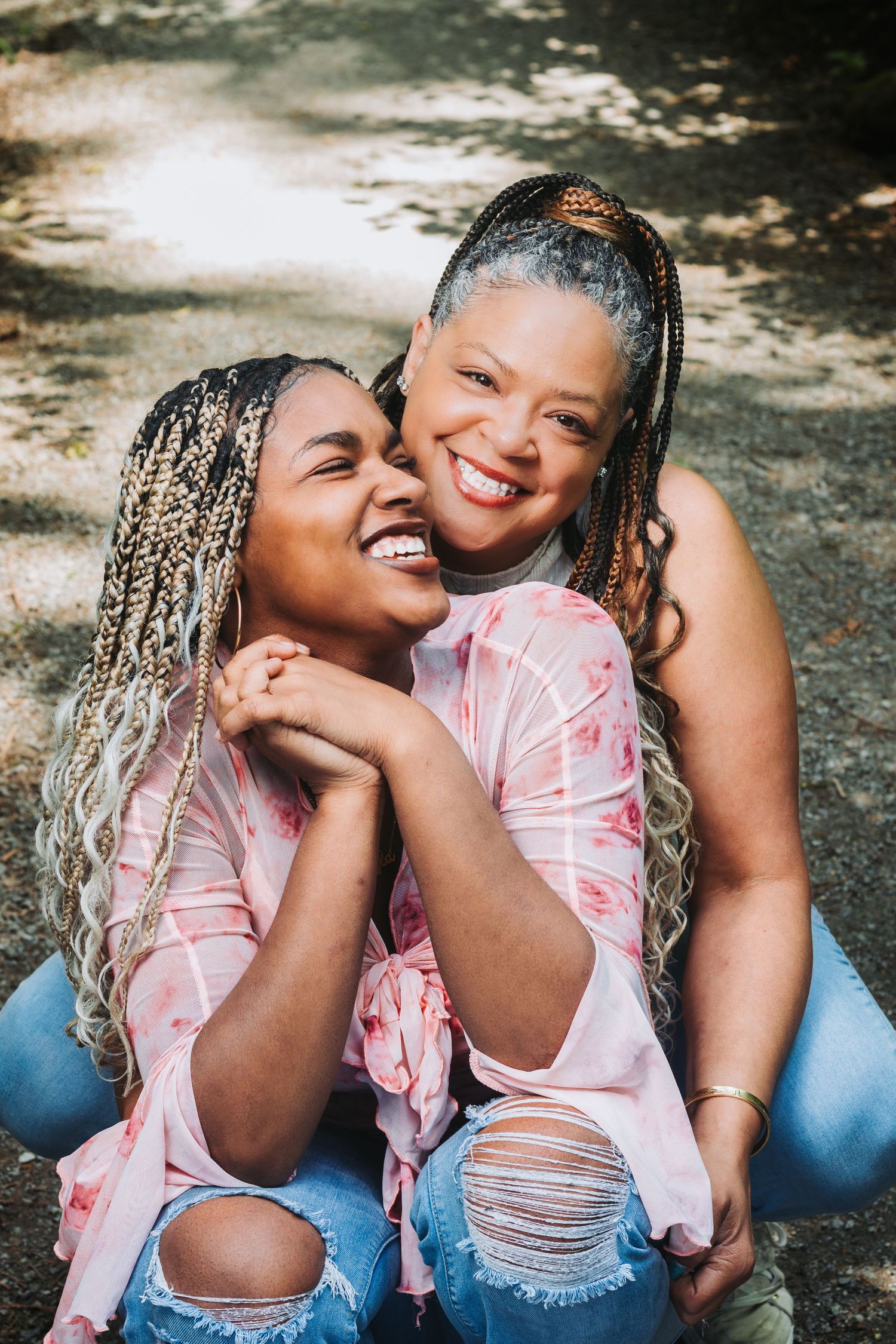 Two women with braids laugh and embrace outdoors, wearing matching pink tops and ripped jeans.