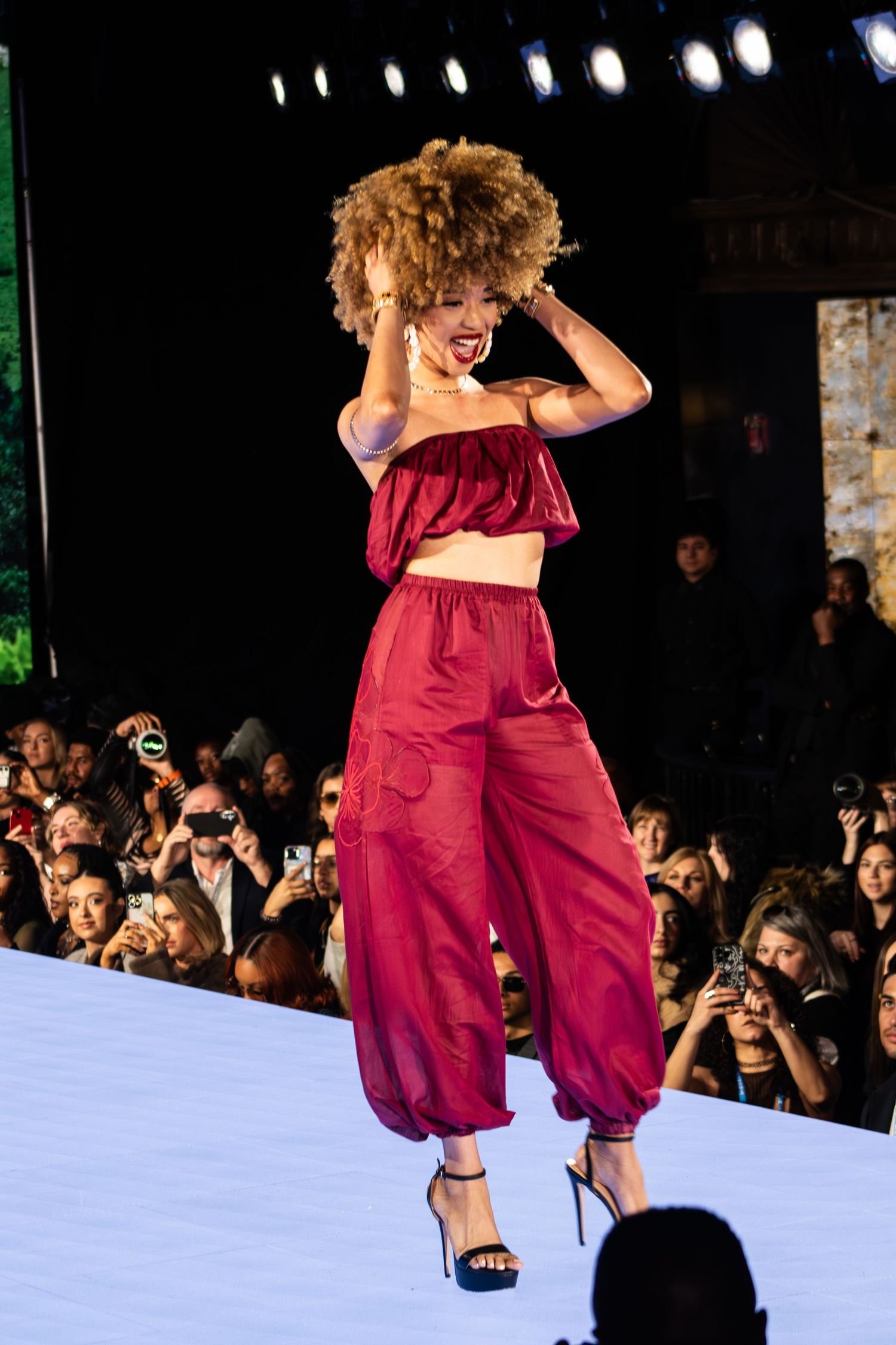Model walks runway in burgundy silk crop top and harem pants ensemble during fashion show performance.