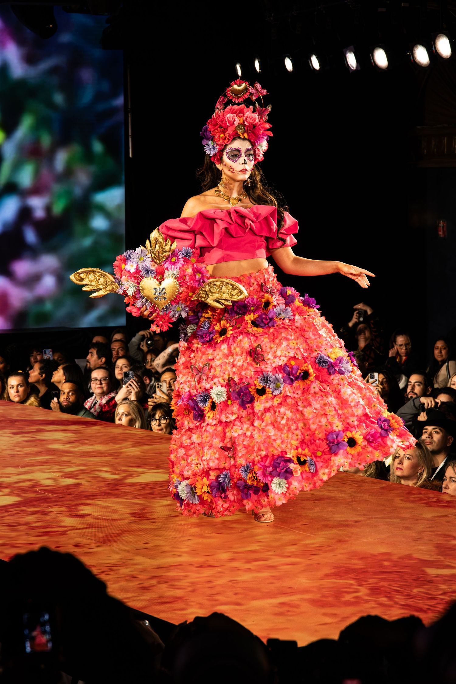A model walks the runway in a vibrant pink floral ball gown with butterfly accents at a fashion show.