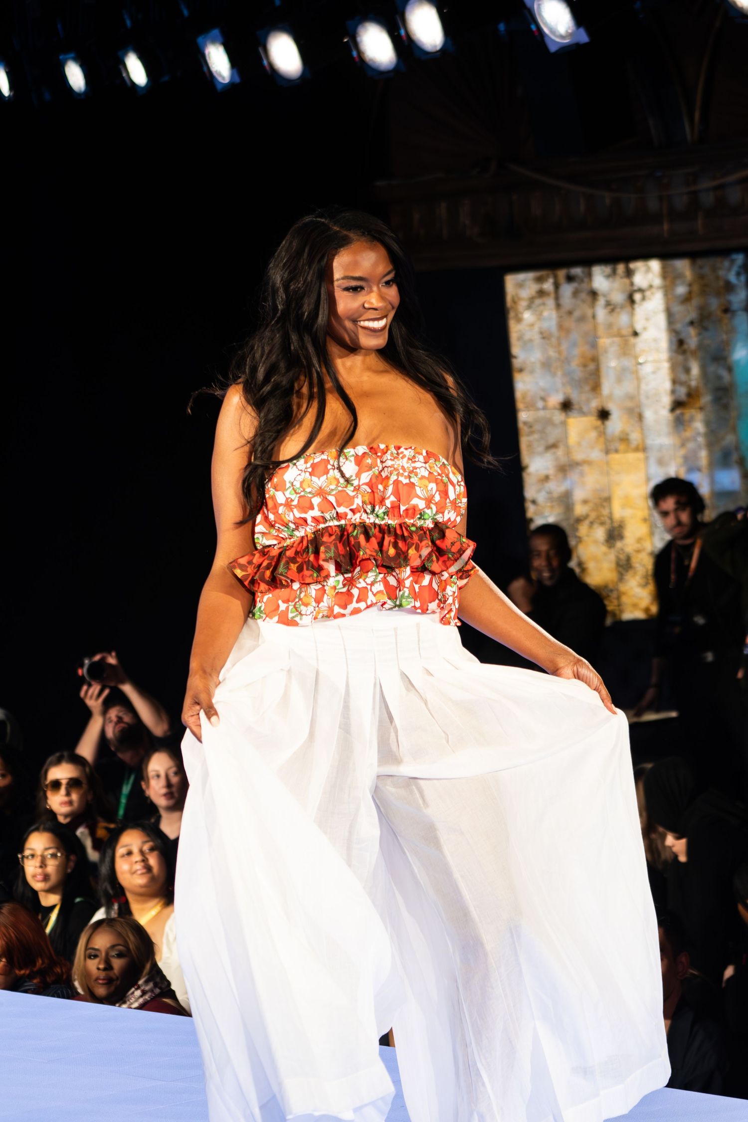 Model walks runway in flowing white maxi skirt and floral strapless top during fashion show.