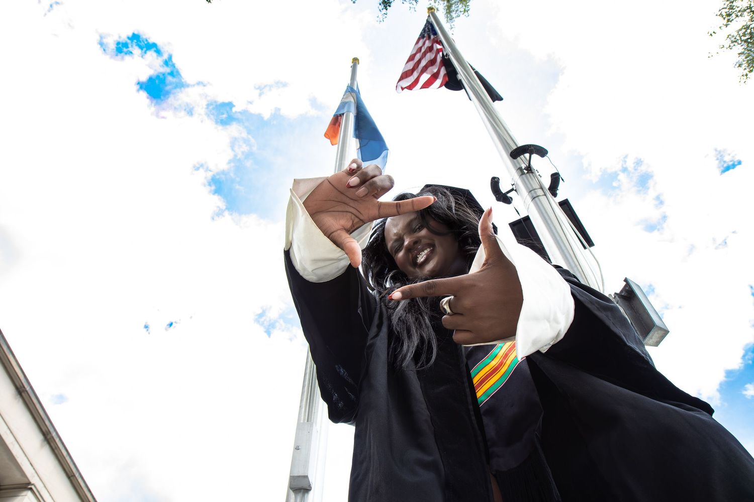 Graduate in cap and gown makes hand gesture beneath American and pride flags against blue sky.