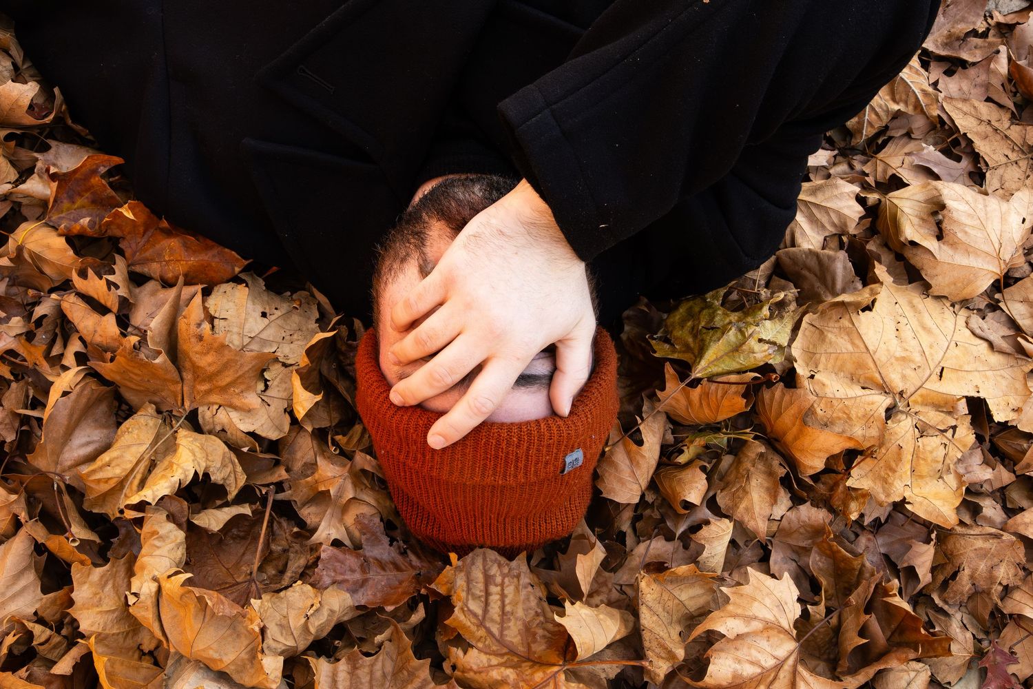Person wearing an orange beanie hat lying among fallen autumn leaves in a forest setting.