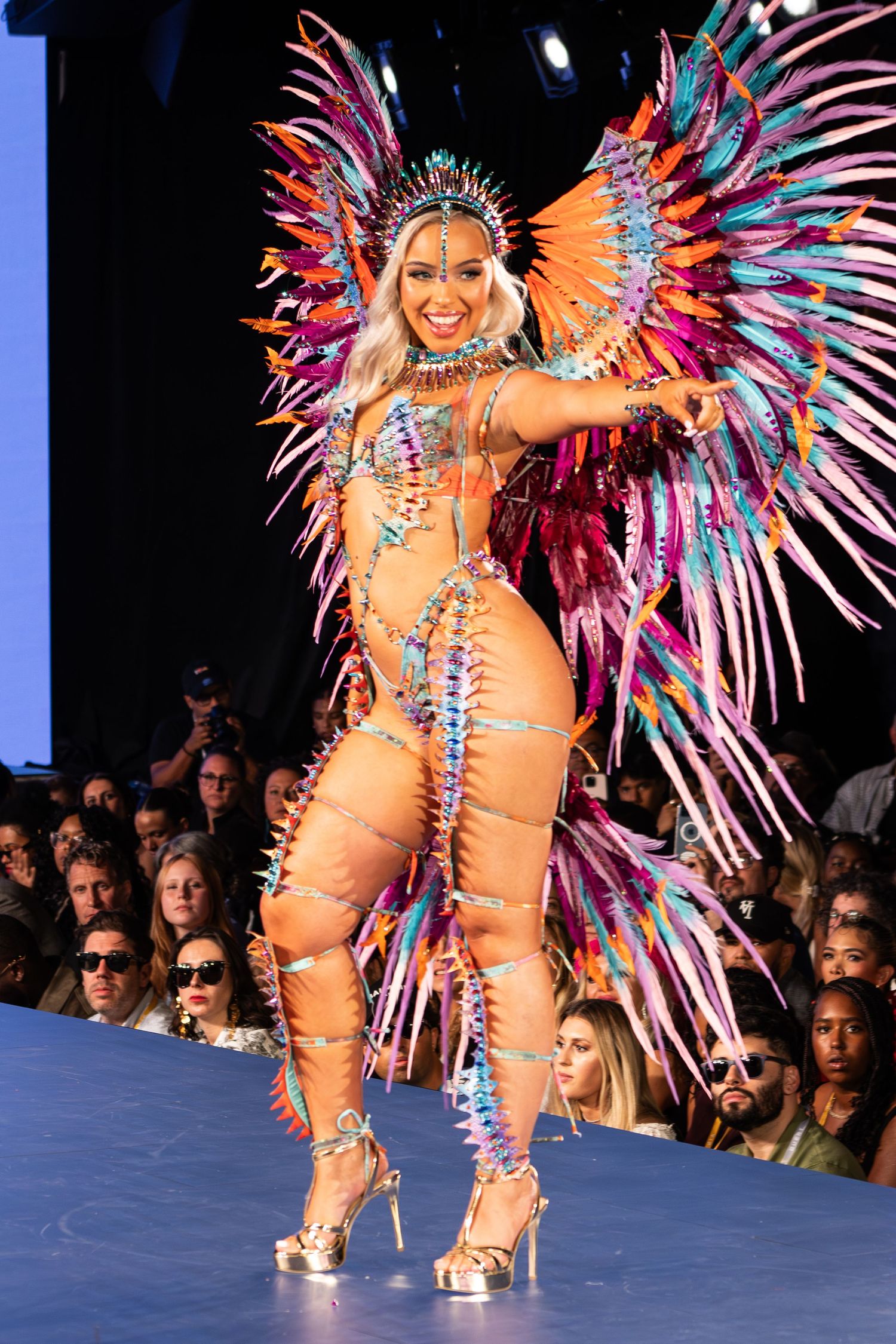 Model wears vibrant carnival costume with colorful feathered wings and beaded bodysuit on fashion runway.