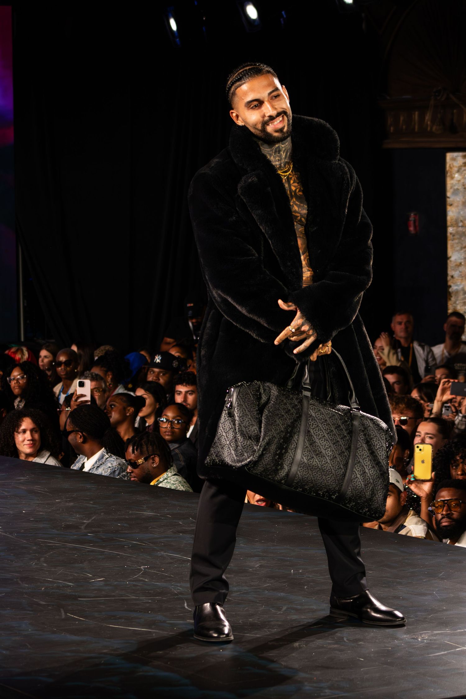 Man in black fur coat holding luxury duffel bag walks runway at fashion show.