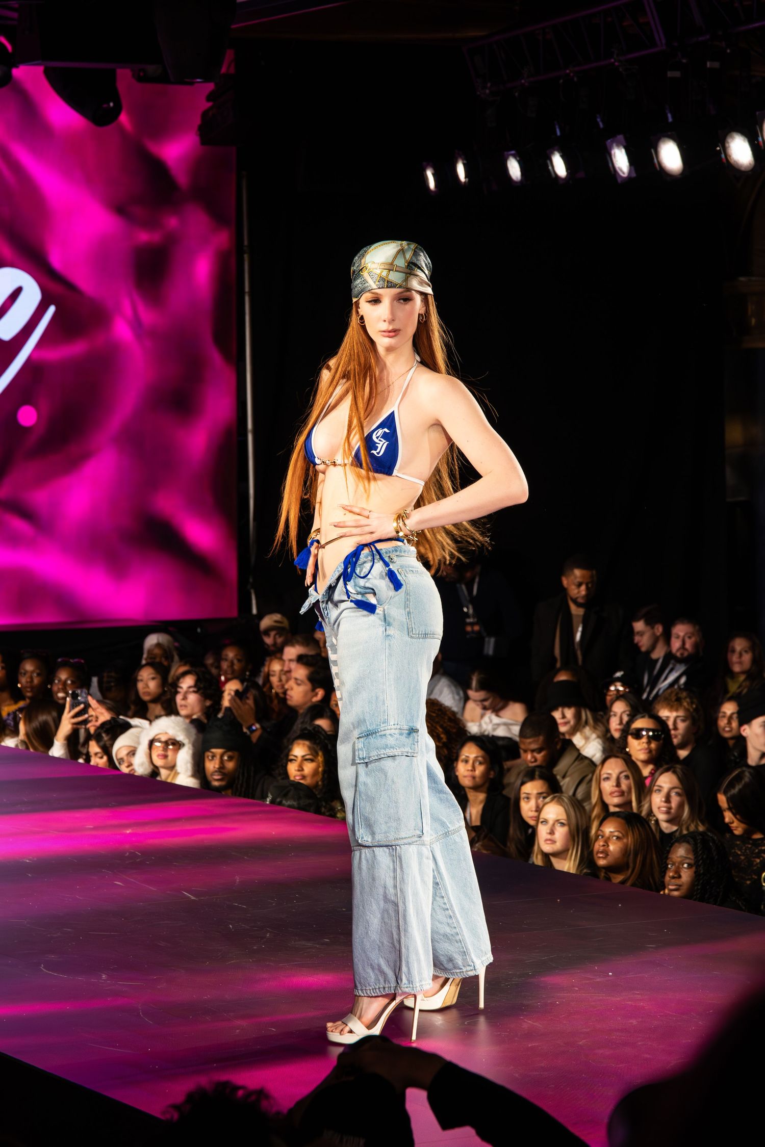 Model walks runway in blue bikini top and light wash cargo jeans at fashion show with pink lighting and audience.