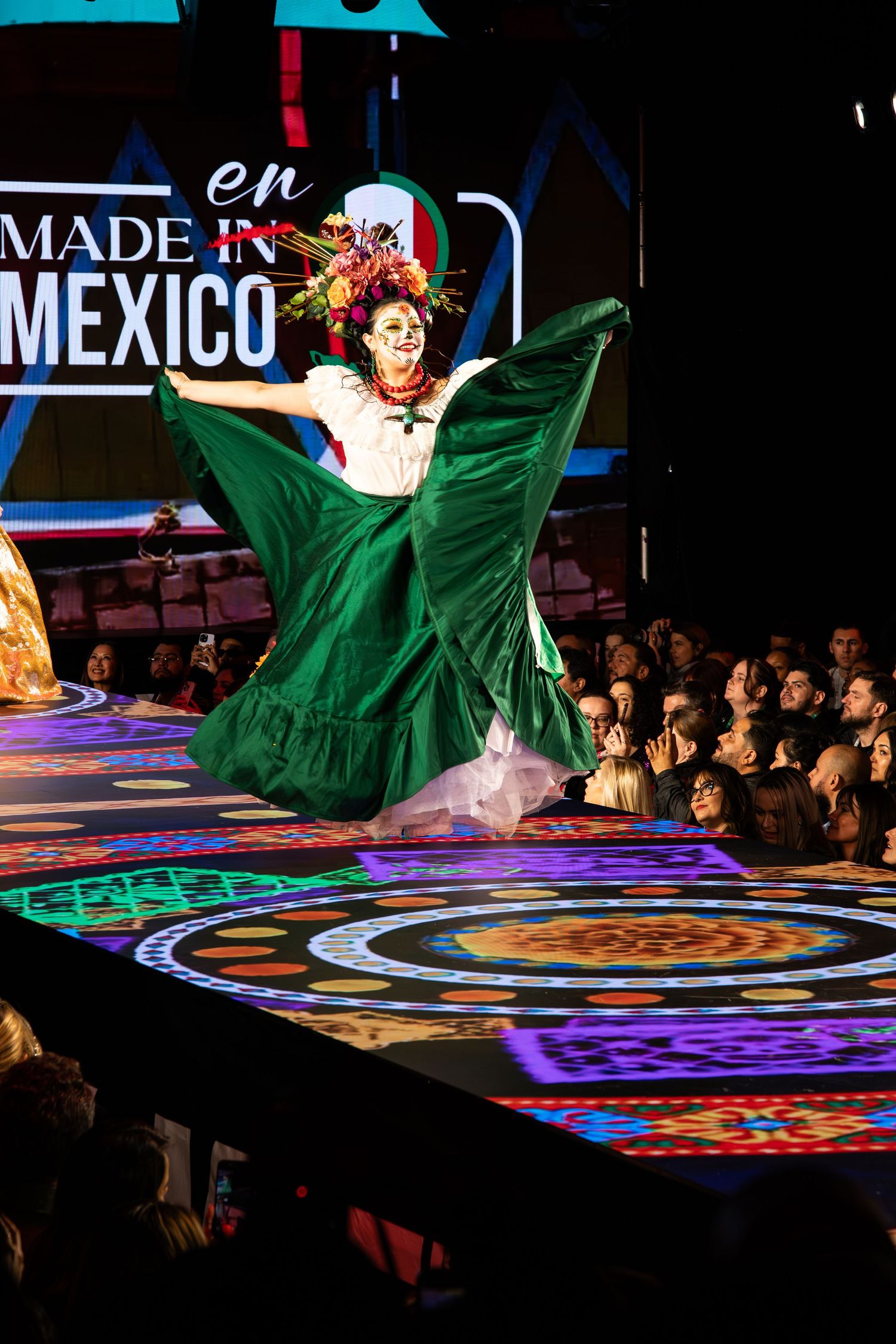 A dancer in a vibrant green traditional Mexican dress performs on an illuminated stage with colorful projected patterns.