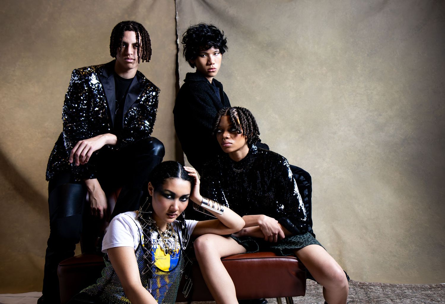 A stylish group poses together in edgy dark outfits with one member wearing a white t-shirt against a neutral backdrop.