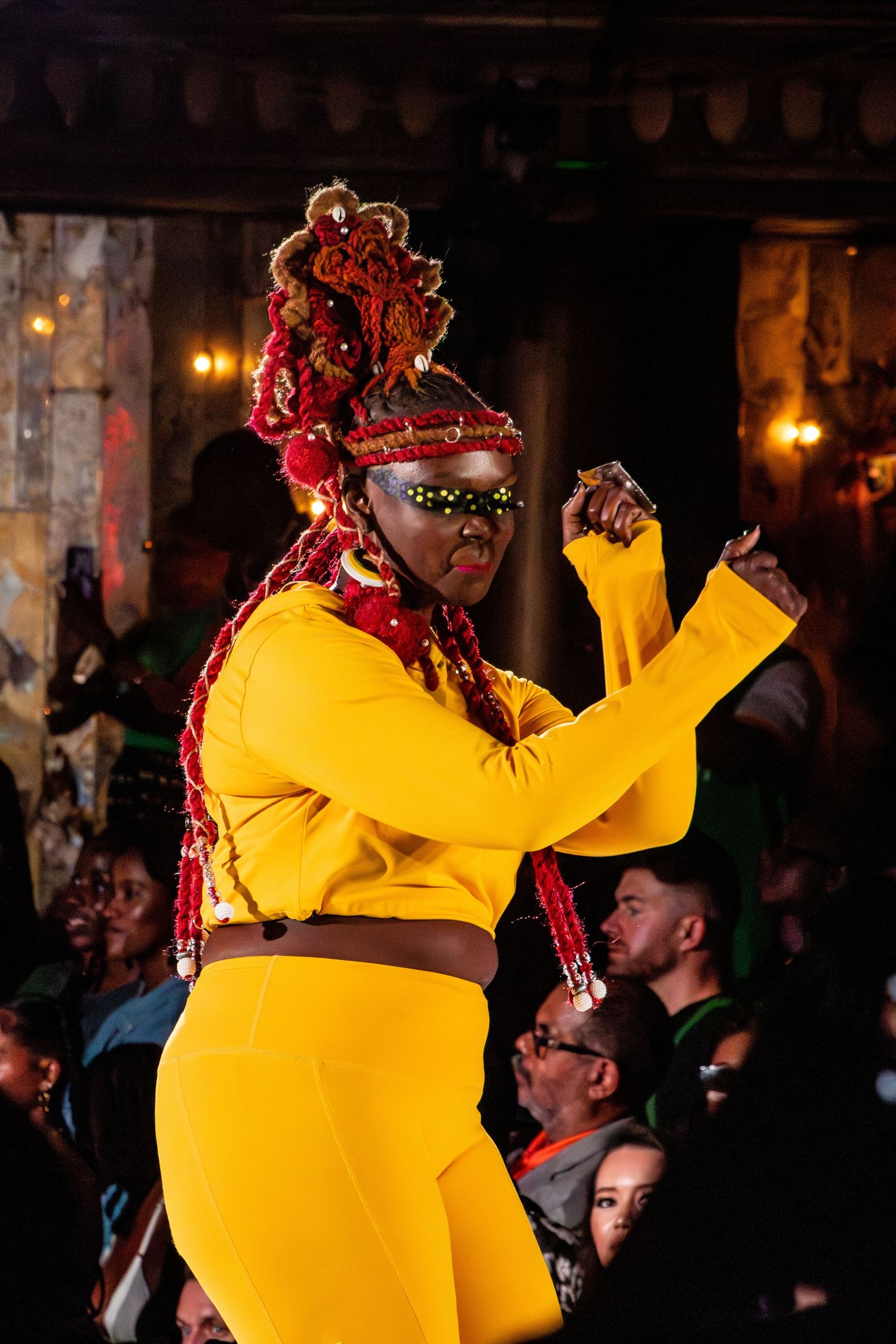 Performer in bright yellow outfit and distinctive hairstyle dances on stage.