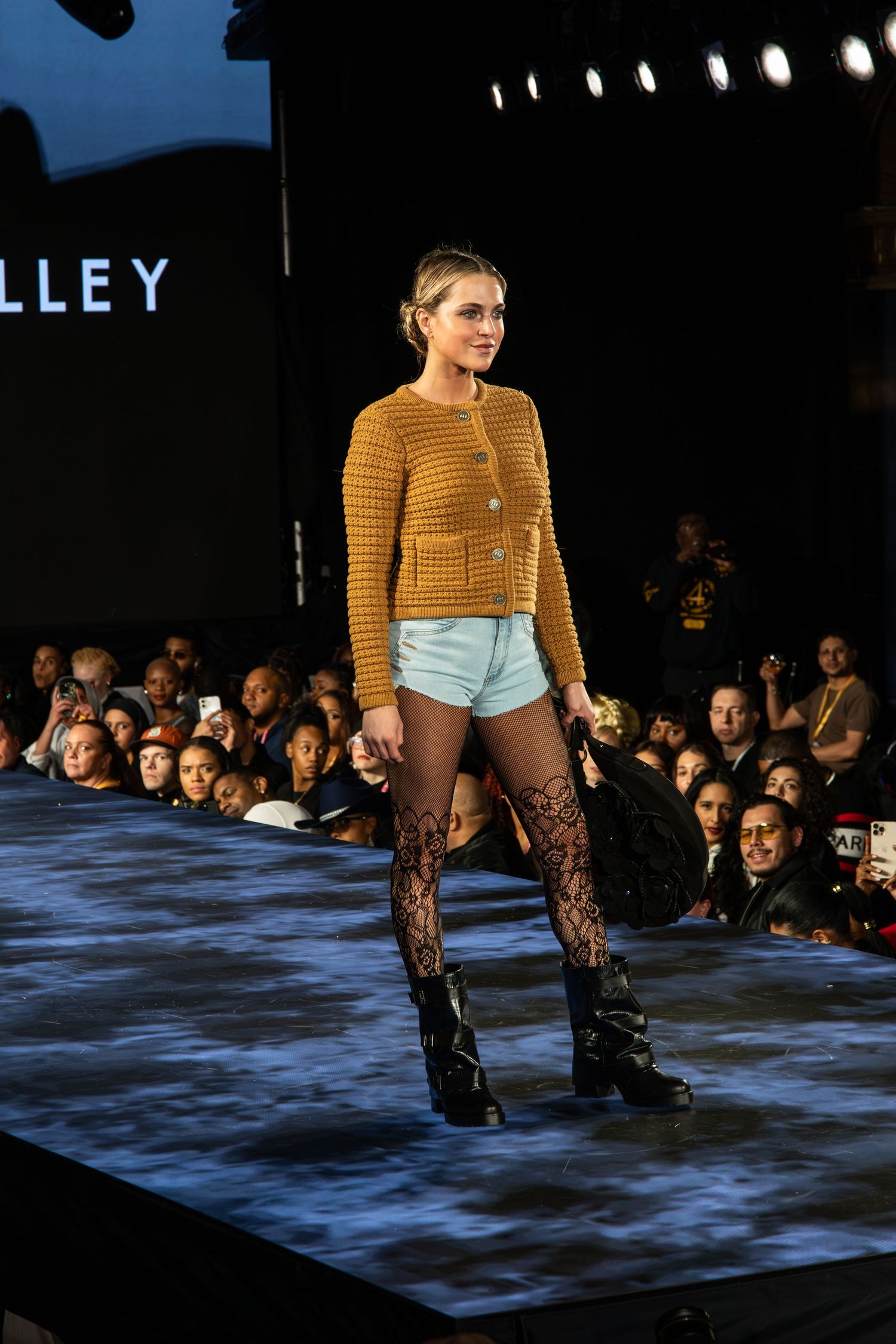 Fashion model walks runway wearing mustard yellow sweater, denim shorts and lace patterned stockings with combat boots.