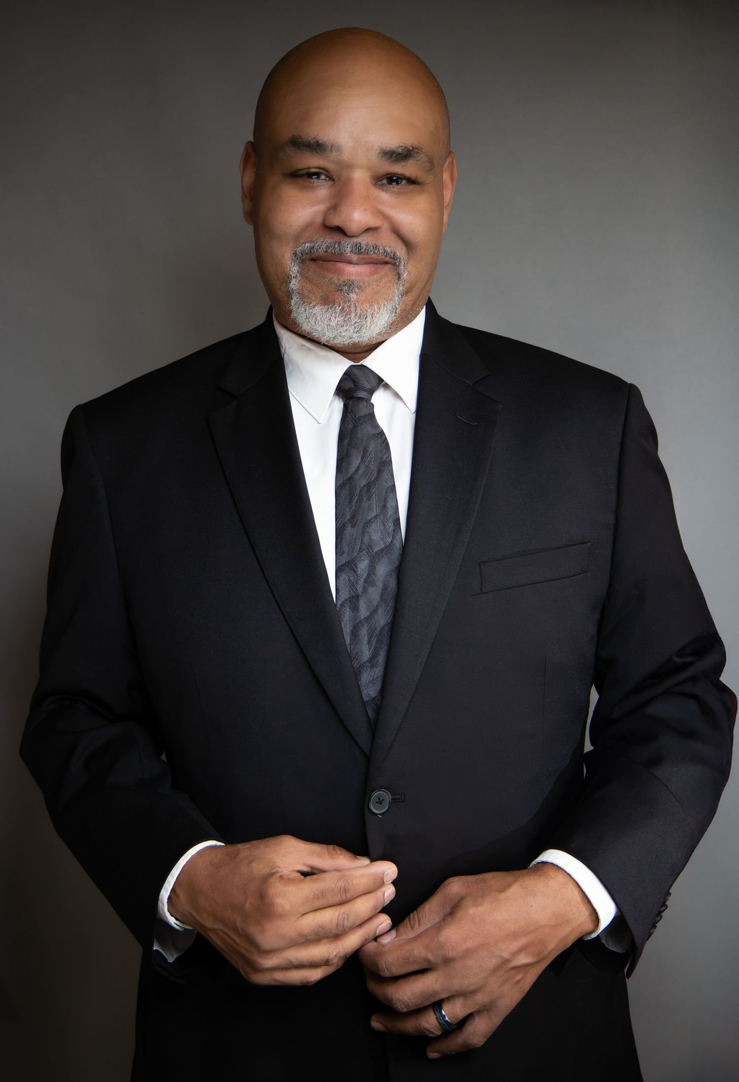 Professional man in black suit and tie posing confidently against a gray background.