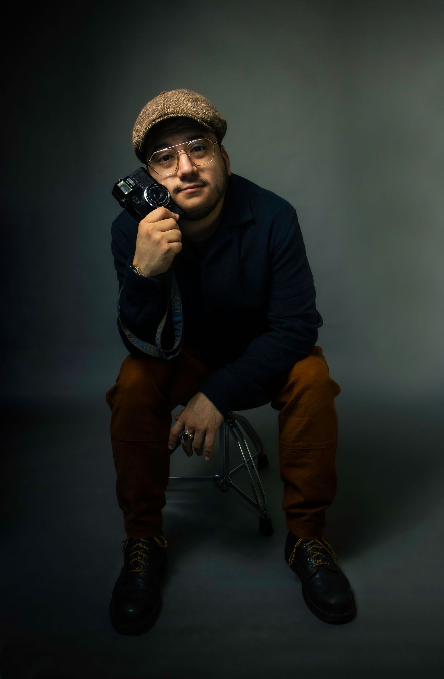 Person in navy sweater and brown pants holding camera while sitting on stool in dark studio.