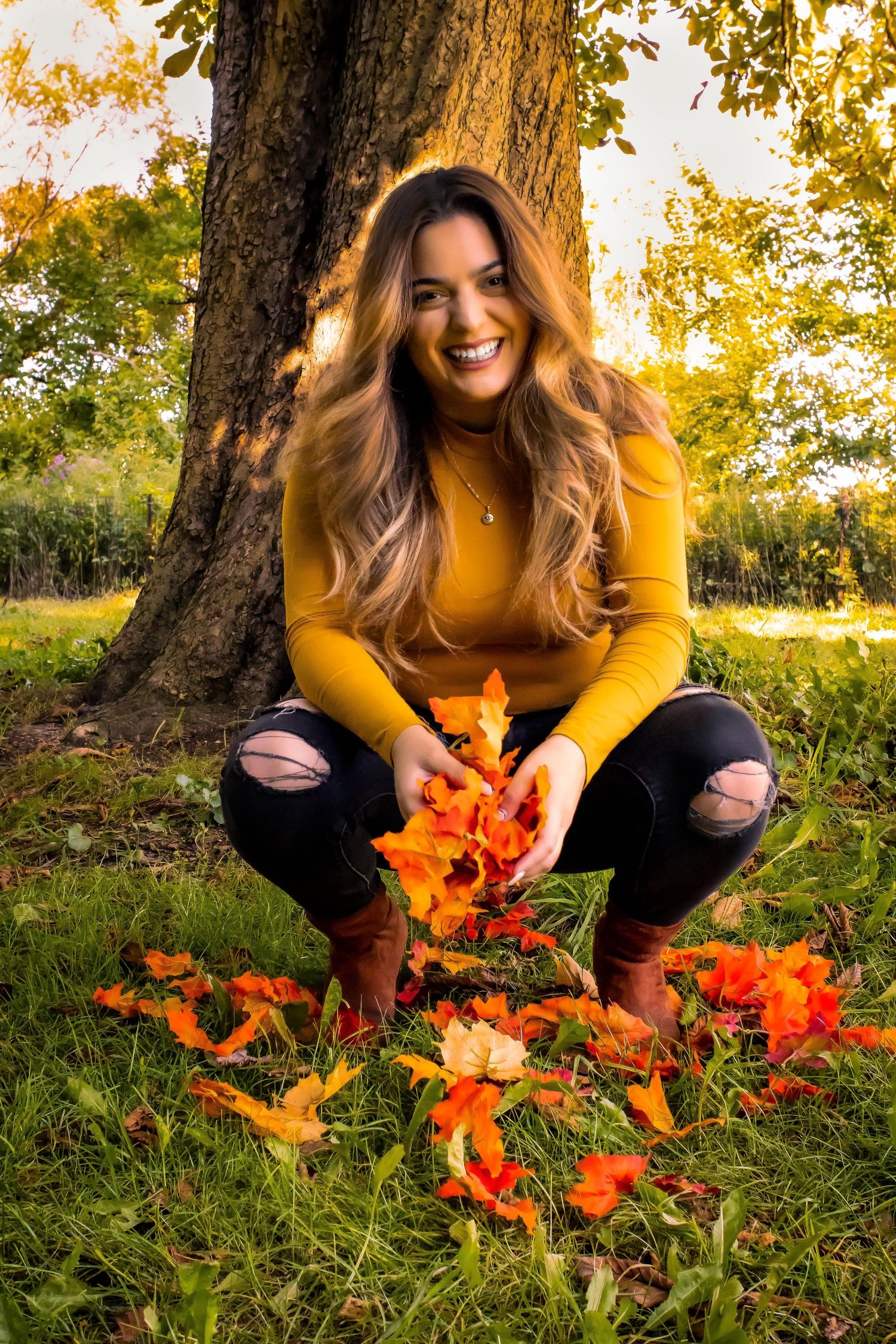 Person in yellow sweater playing with orange autumn leaves under a tree at sunset.