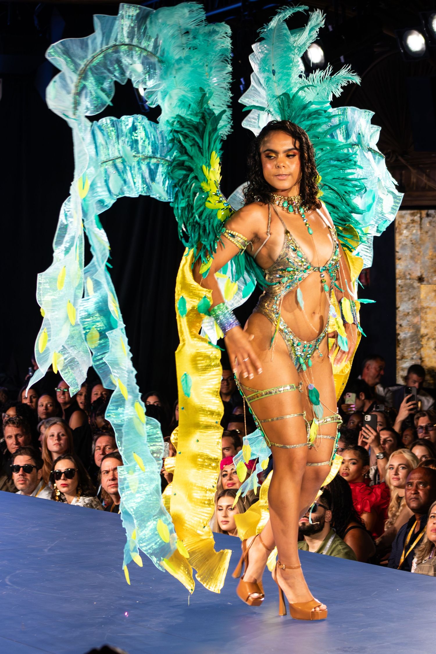 Model walks runway in teal feathered carnival costume with yellow ruffles at fashion show.
