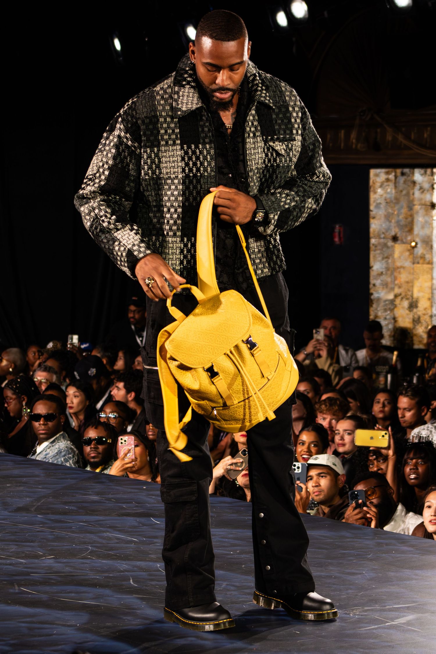 Male model examines yellow backpack on runway wearing dark patterned jacket and black pants at fashion show.