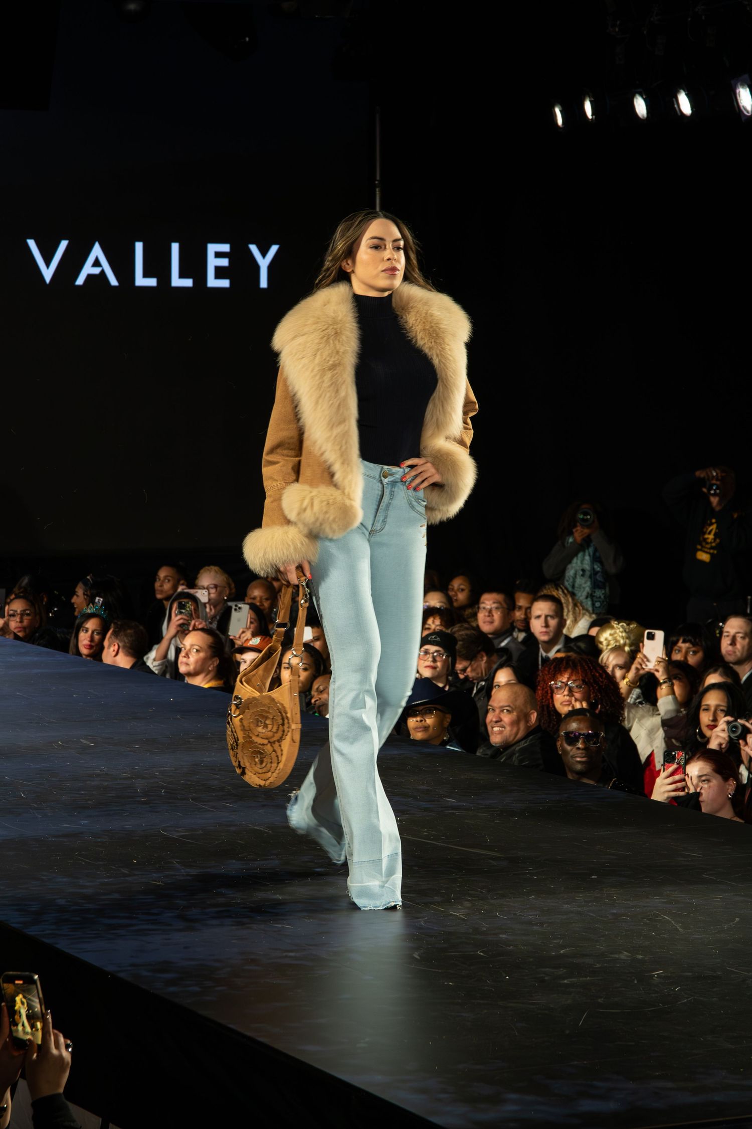 Model walks runway at fashion show wearing tan fur-trimmed coat, light jeans and carrying round handbag on dark catwalk.