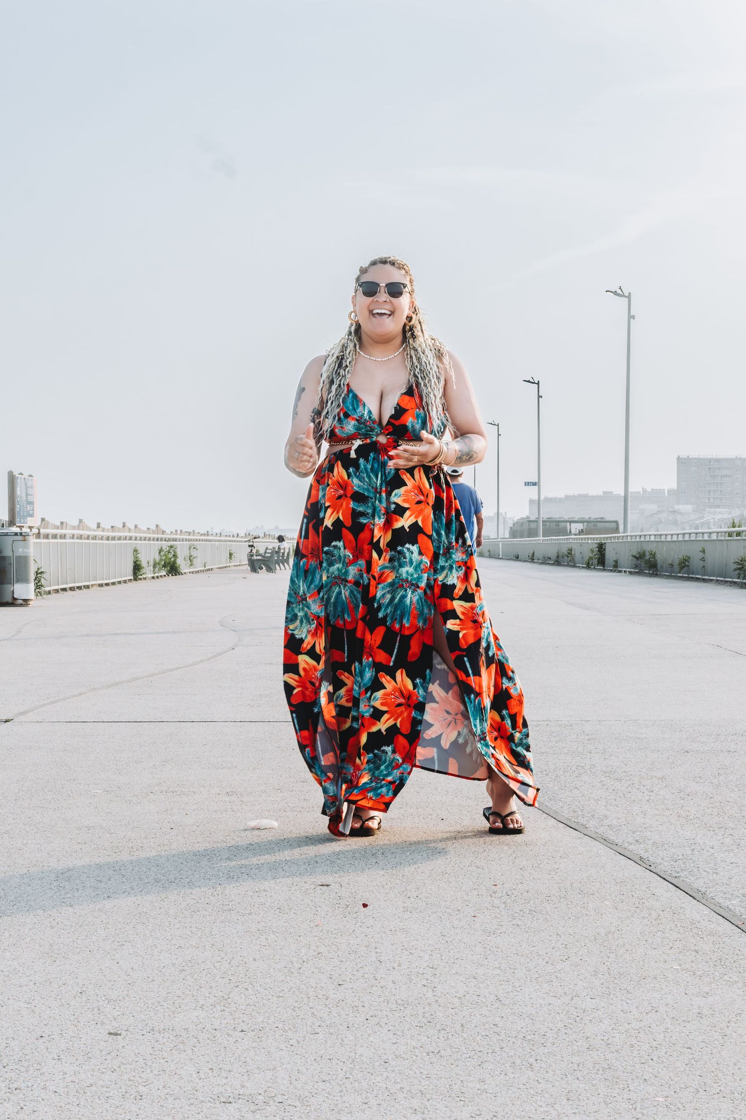 Woman in colorful floral maxi dress and sunglasses walking on urban bridge on a sunny day.