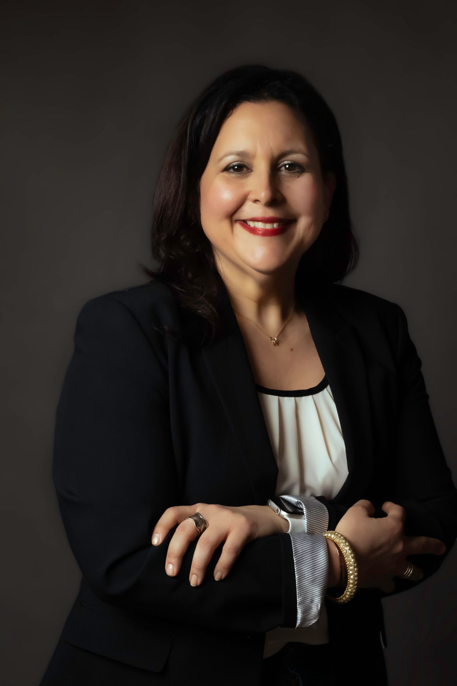 Professional studio portrait of someone in business attire with jewelry against dark background.