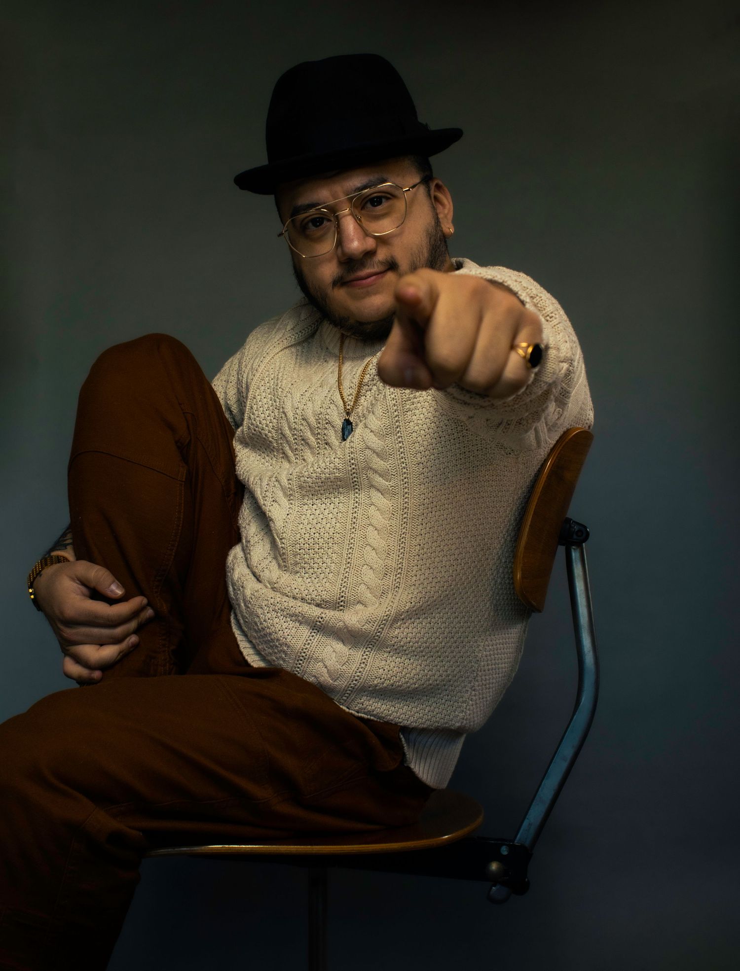 Studio portrait of someone in a cream cable knit sweater and fedora pointing at the camera in dramatic lighting.
