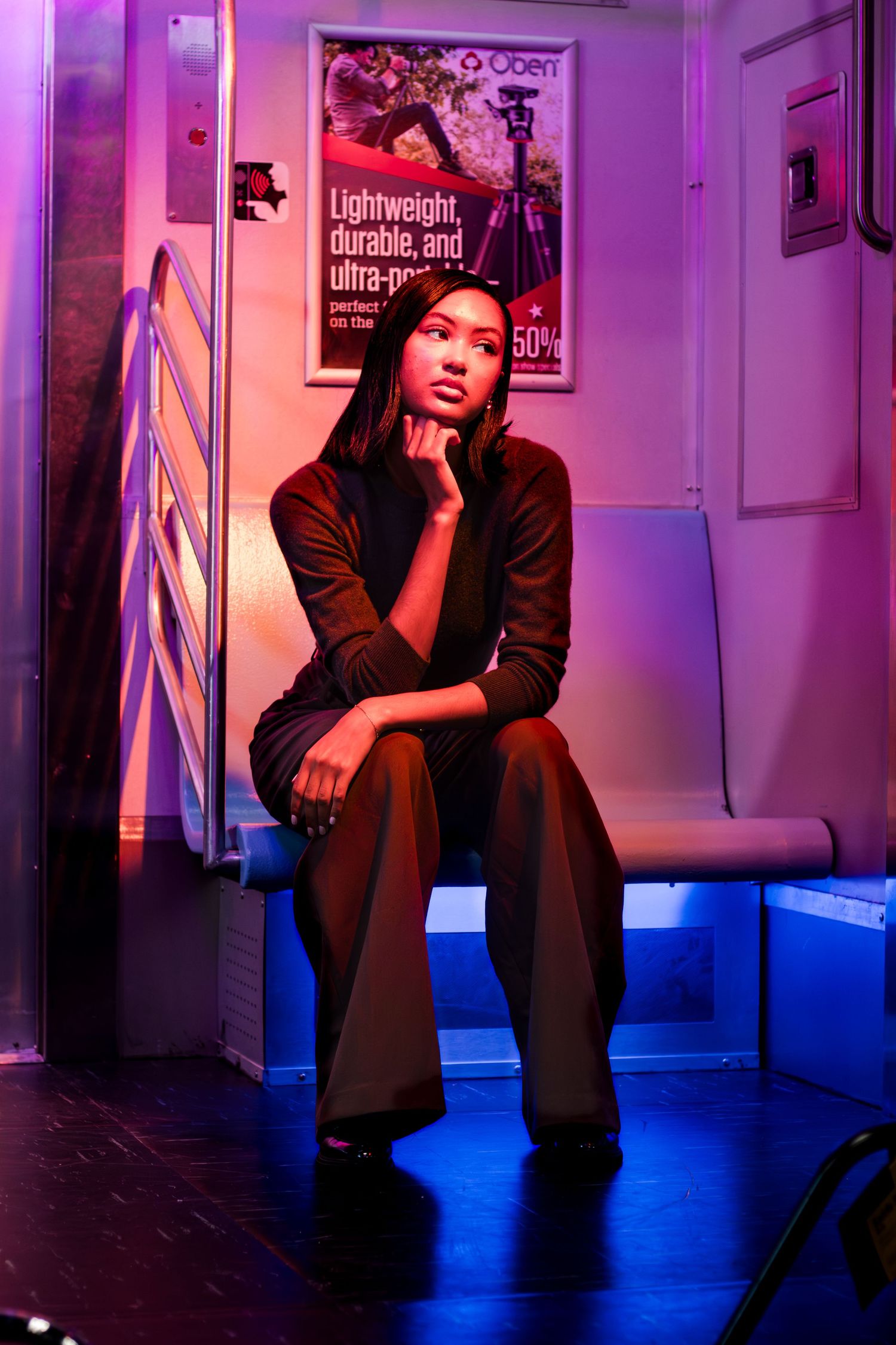 A stylish woman in dark clothing sits pensively on a subway seat bathed in neon pink and blue lights.