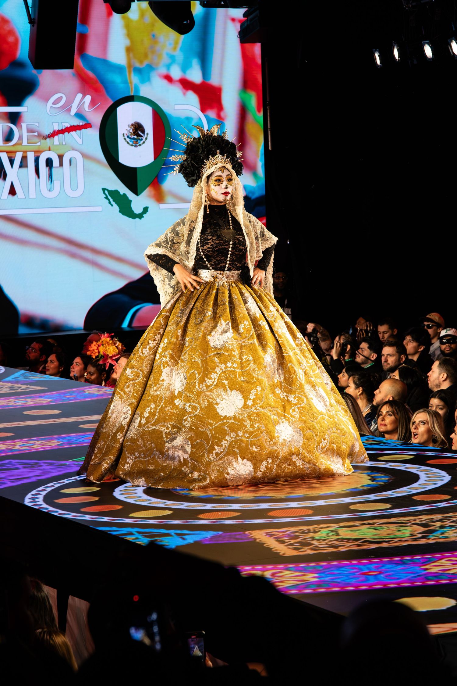 Fashion model walks runway in ornate gold Mexican-inspired ball gown with traditional headpiece at cultural fashion show.